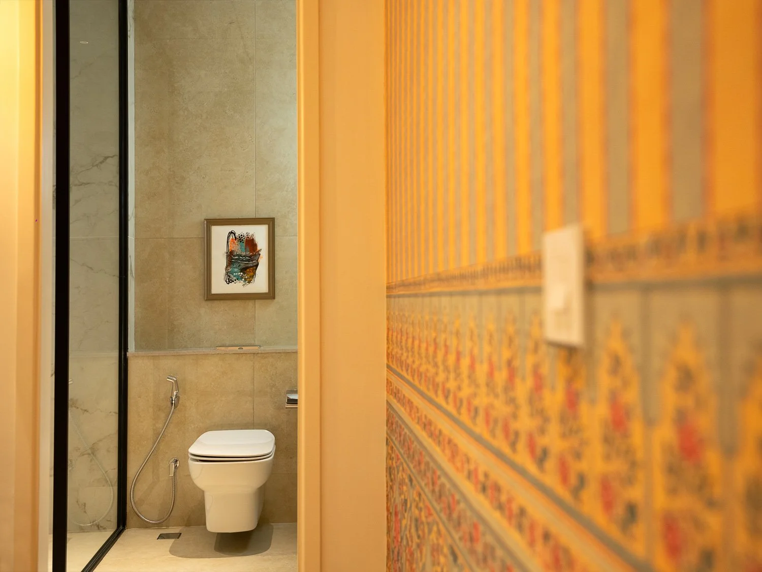 A glimpse into a bathroom with beige tile walls, a modern white toilet, a bidet spray, and a framed abstract painting hanging on the wall. The photo is taken from a hallway with orange and gold patterned wallpaper.