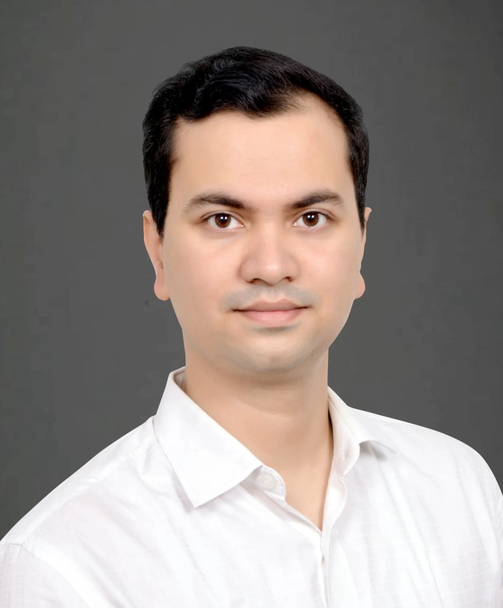 A smiling young woman with curly afro hair wearing glasses, hoop earrings, and layered necklaces, in a cozy indoor setting.