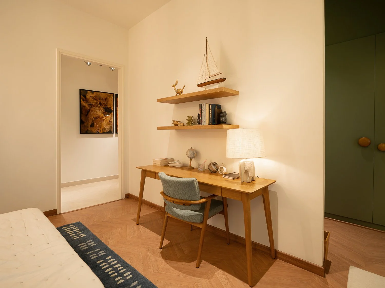 A cozy bedroom corner with a wooden desk, a gray upholstered chair, and floating wooden shelves holding decorative items like books, a model sailboat, and ornaments. There's a table lamp on the desk and a patterned rug on the wooden floor.