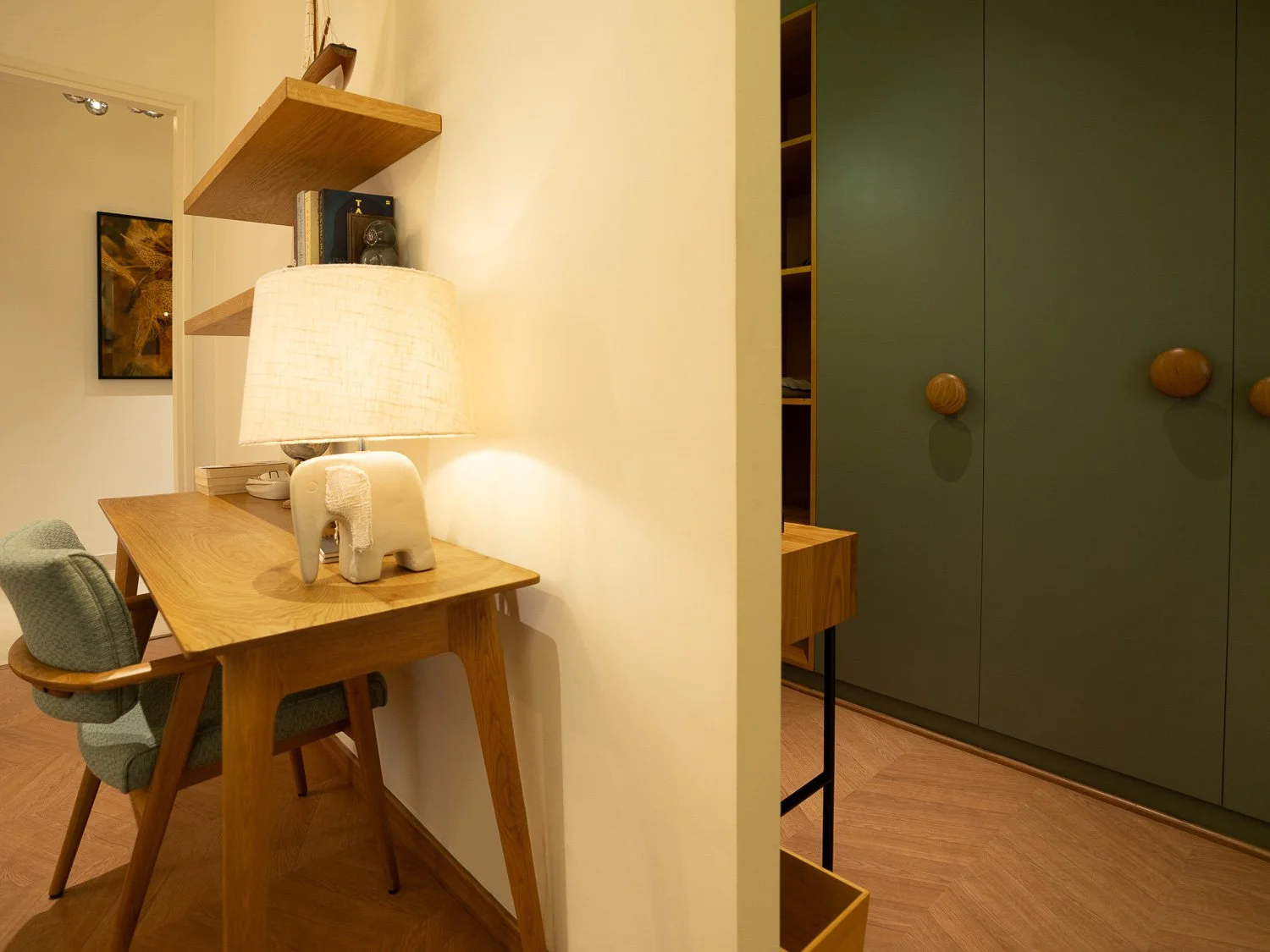 Interior view of a room with wooden furniture, a lamp, a decorative elephant figurine, and shelves with books on the left side, next to a green closet with round wooden handles on the right side.