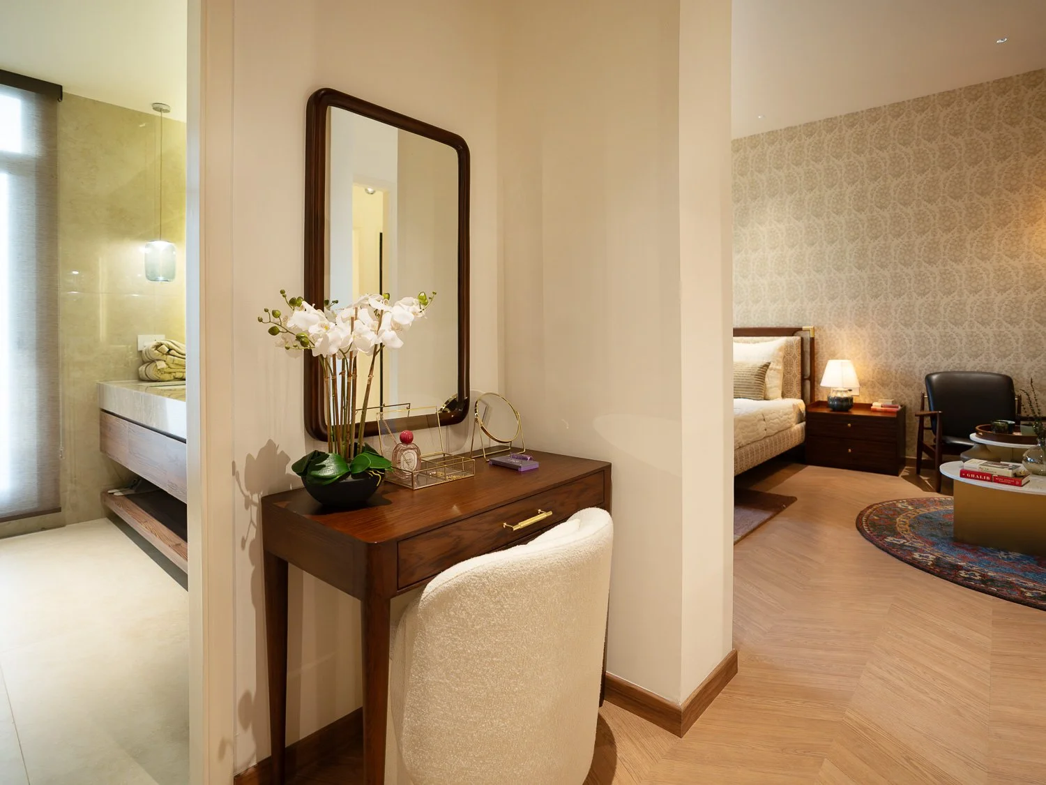 A bedroom with a nightstand, a lamp, and a rug, visible through an open door from a small dressing area with a wooden vanity, mirror, and a white upholstered chair.