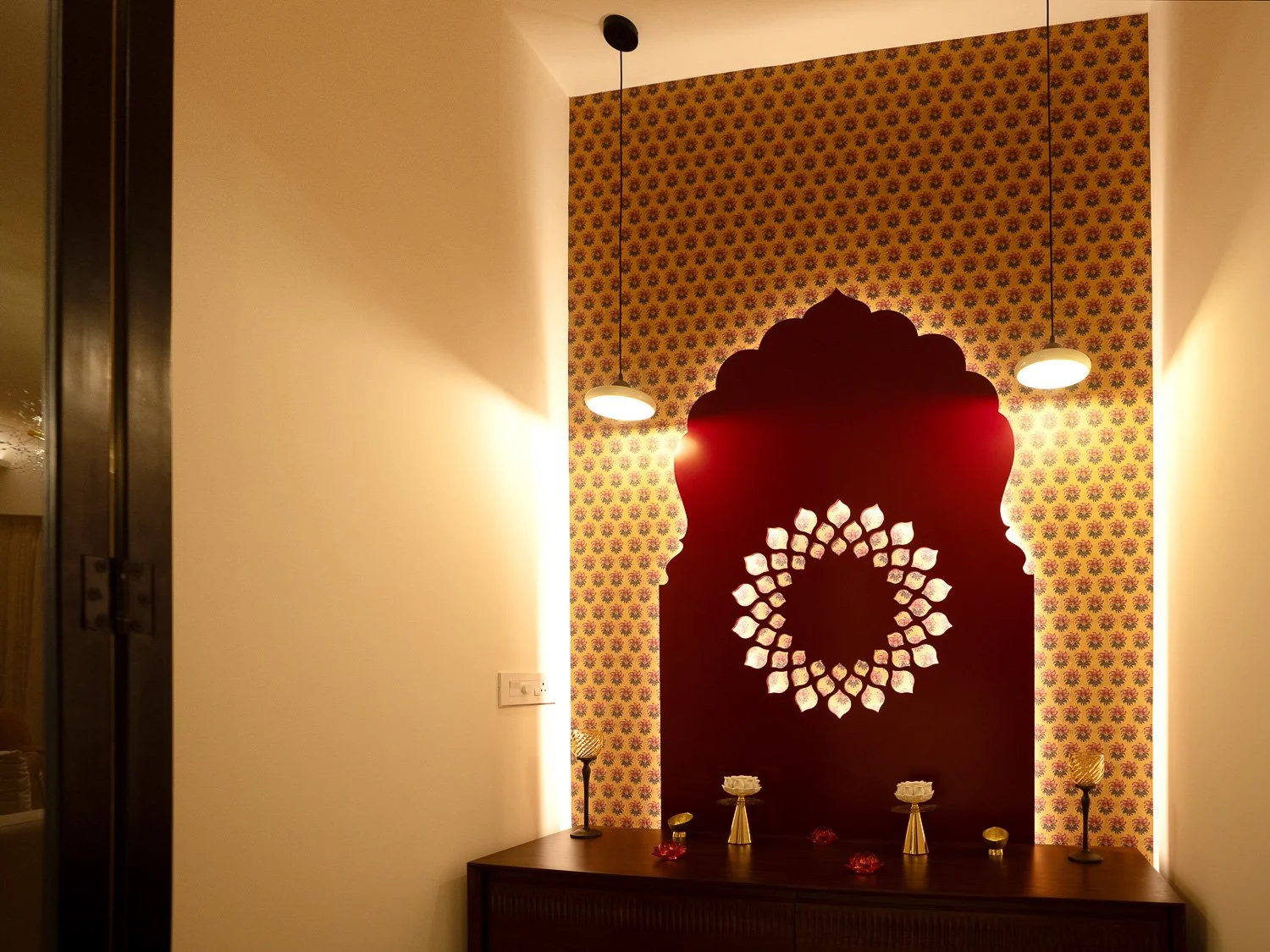 Decorative interior wall with traditional Indian design elements, featuring a large cut-out pattern in a dark red panel, backlit with warm lighting from hanging pendant lights, and a wooden console table with ornamental items.