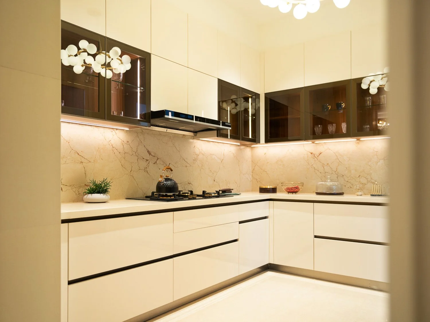 Modern kitchen with cream-colored cabinets, beige marble backsplash, black glass-front upper cabinets, and built-in oven. Items include a kettle, potted plant, glassware, and decorative bowls.