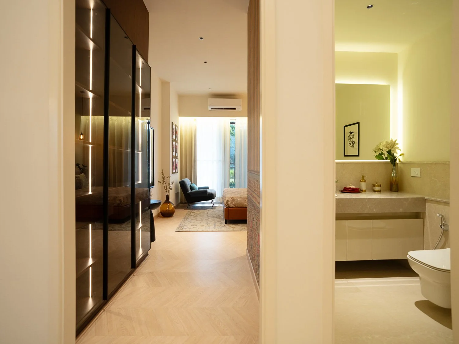 Modern bedroom and bathroom with neutral tones and minimalist decor, featuring a black wardrobe, a framed picture on the wall, a chair, a bed, a vase with flowers, and a vanity.