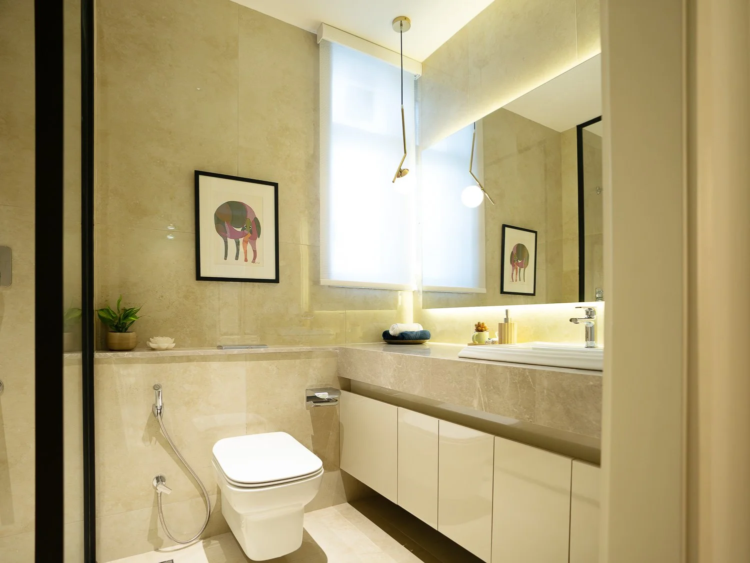 Modern bathroom with beige marble walls and countertop, white cabinetry, a window with frosted glass, framed colorful art, and minimal decor.