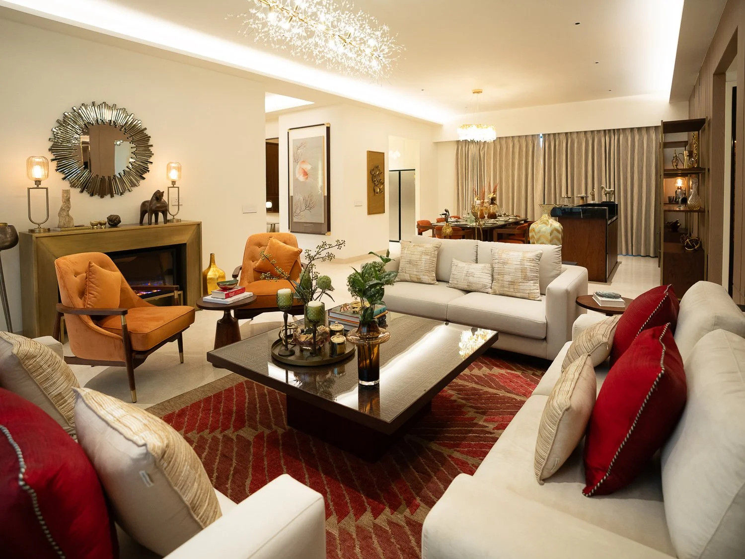 Living room with white sofas, red and beige pillows, orange armchairs, a dark coffee table, and a red patterned rug. There are decorative items on the table and shelves, a fireplace with a large mirror above, and a dining area in the background.