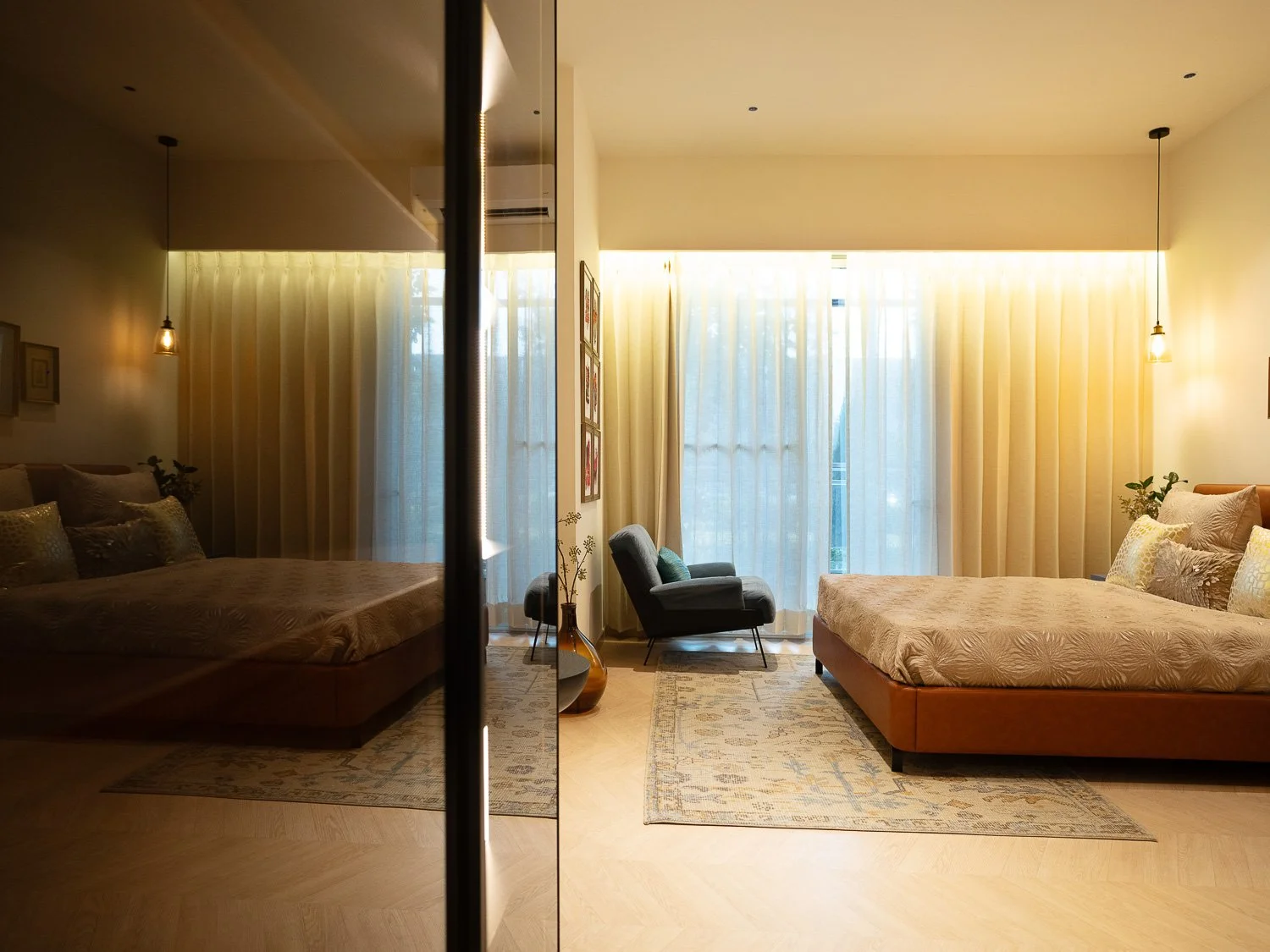 A modern bedroom with large windows covered by sheer curtains, featuring two beds with pillows, a gray armchair, and a tall decorative vase, with soft warm lighting.