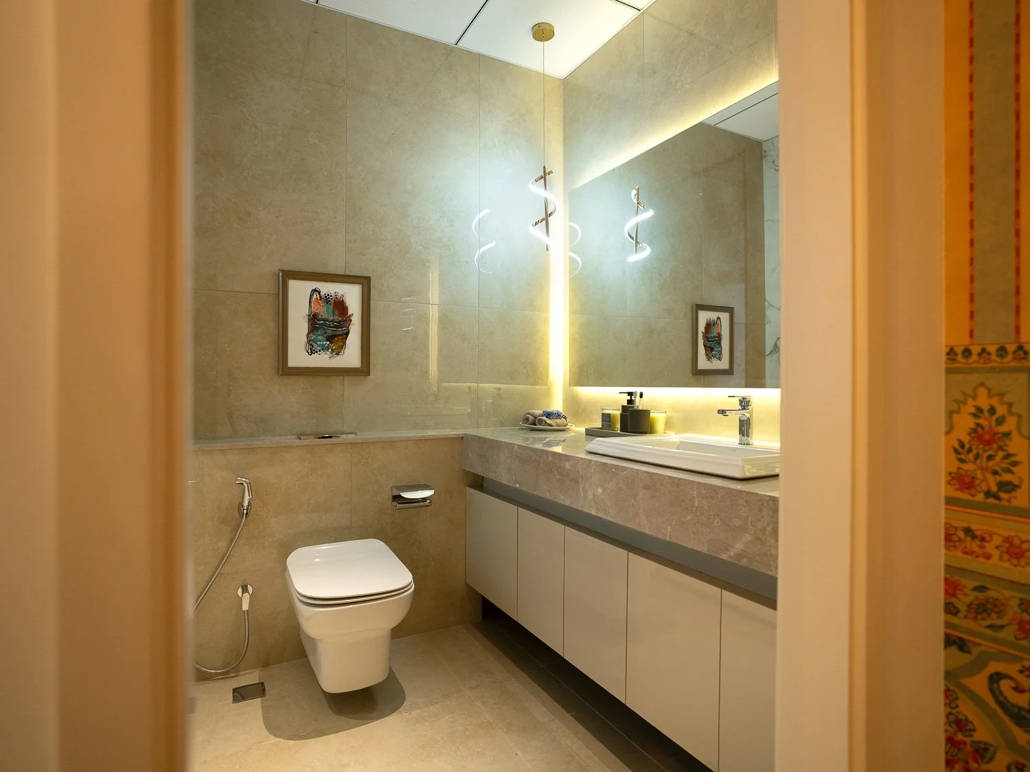 Modern bathroom with beige tiles, a large mirror with LED lighting, a white sink with a gray countertop, and a wall-mounted toilet with a bidet hose. Artwork hangs on the wall beside the mirror.