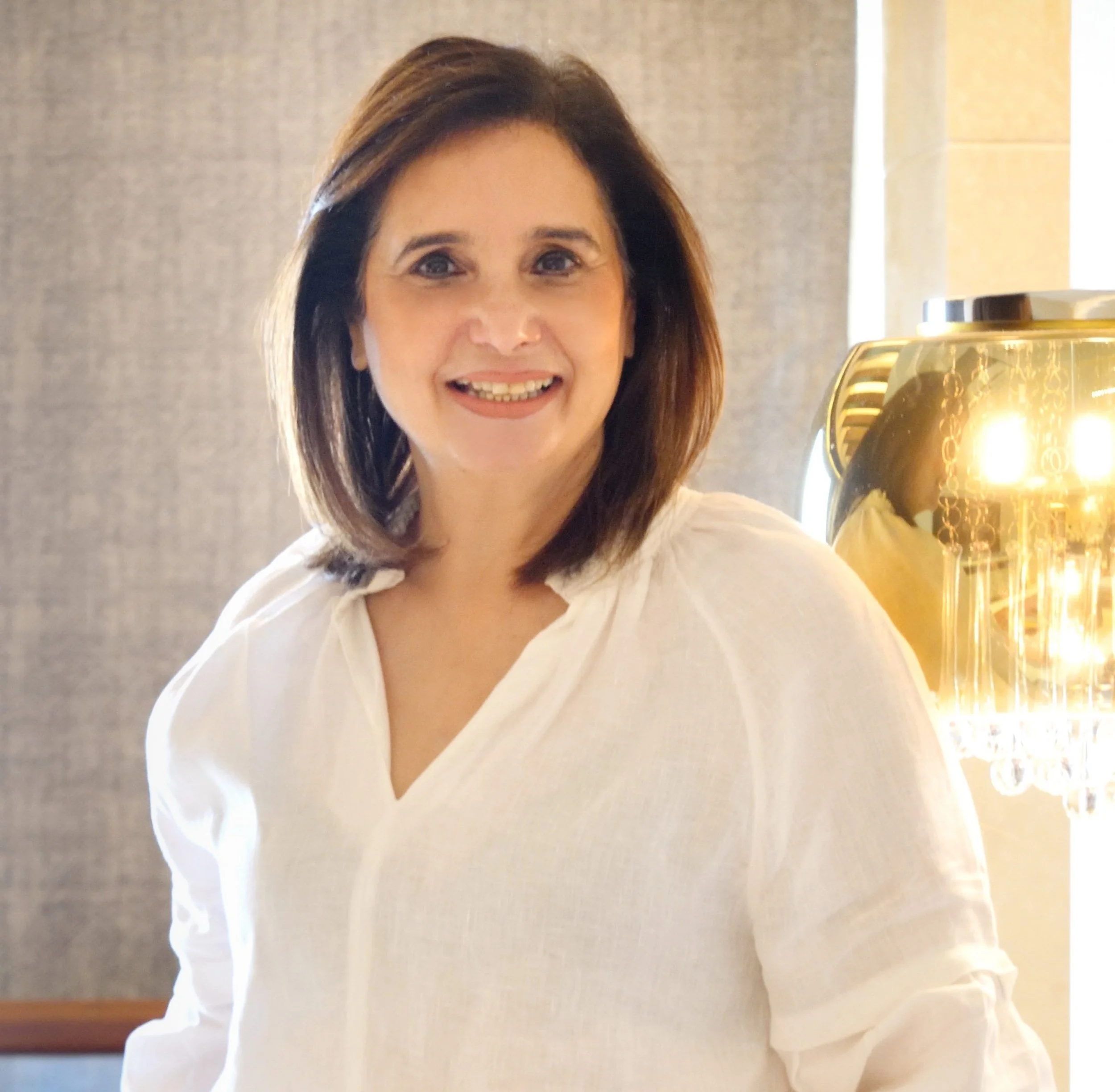 A man with curly dark hair, glasses, and a beard, wearing a dark green sweater, looking thoughtfully to the side in a bright room with a plant in the background.