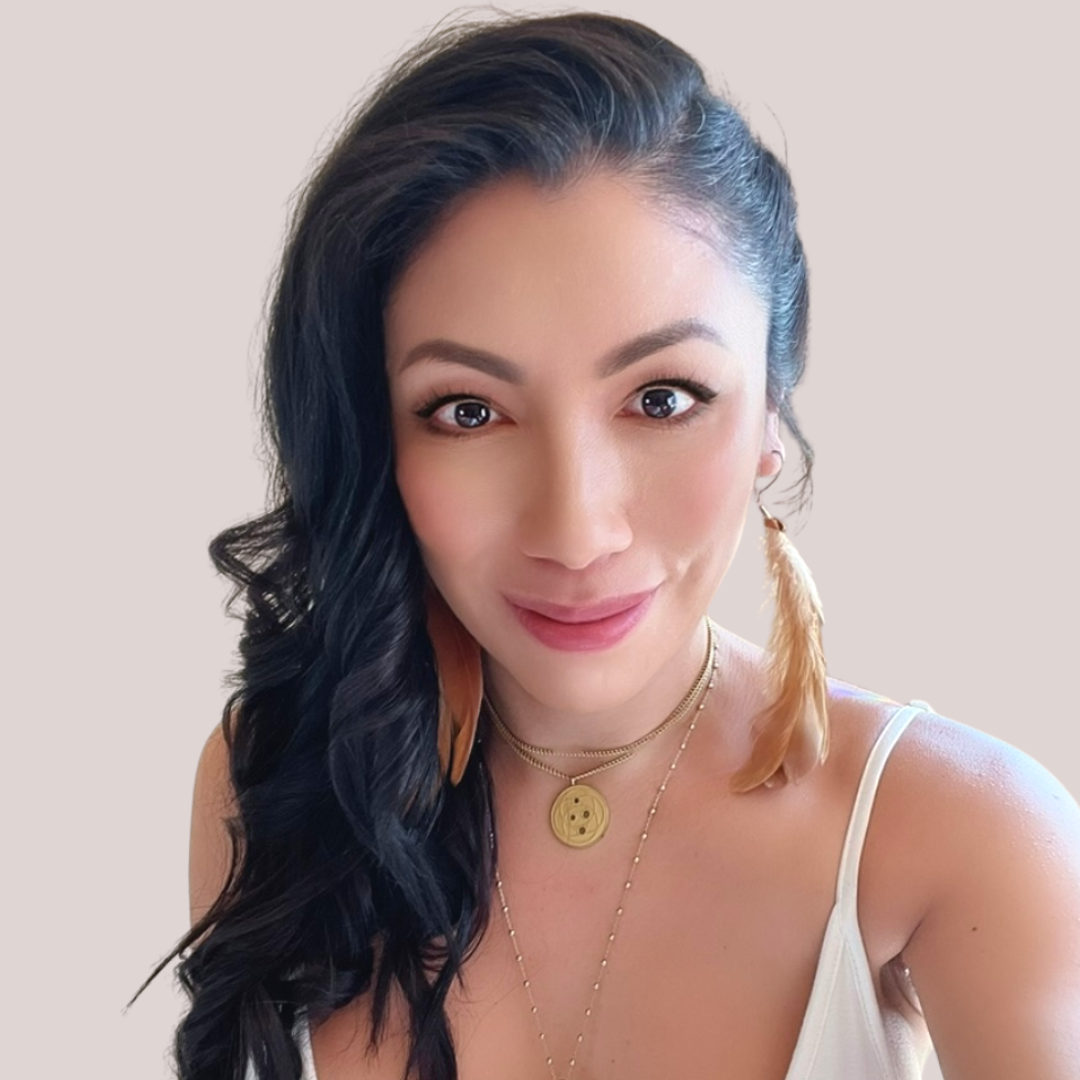 Portrait of a woman with long black hair, wearing a white top, layered gold necklaces, and feather earrings, smiling at the camera.