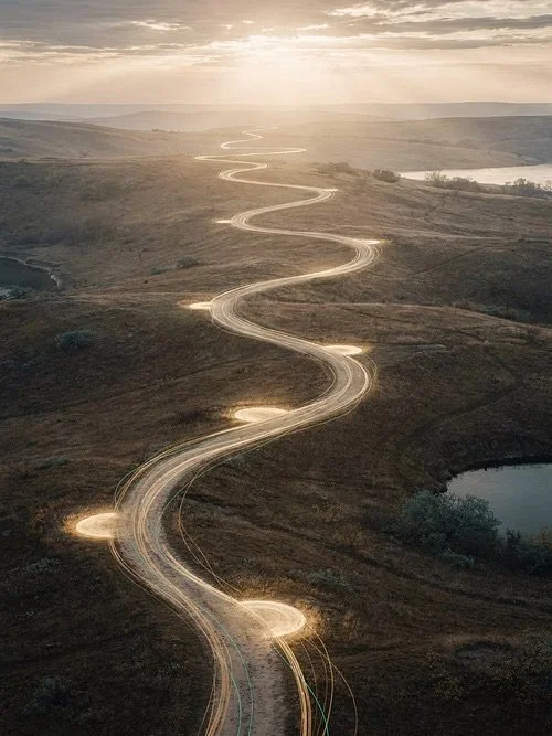 A winding dirt road across a hilly landscape during sunset, with car light trails visible.