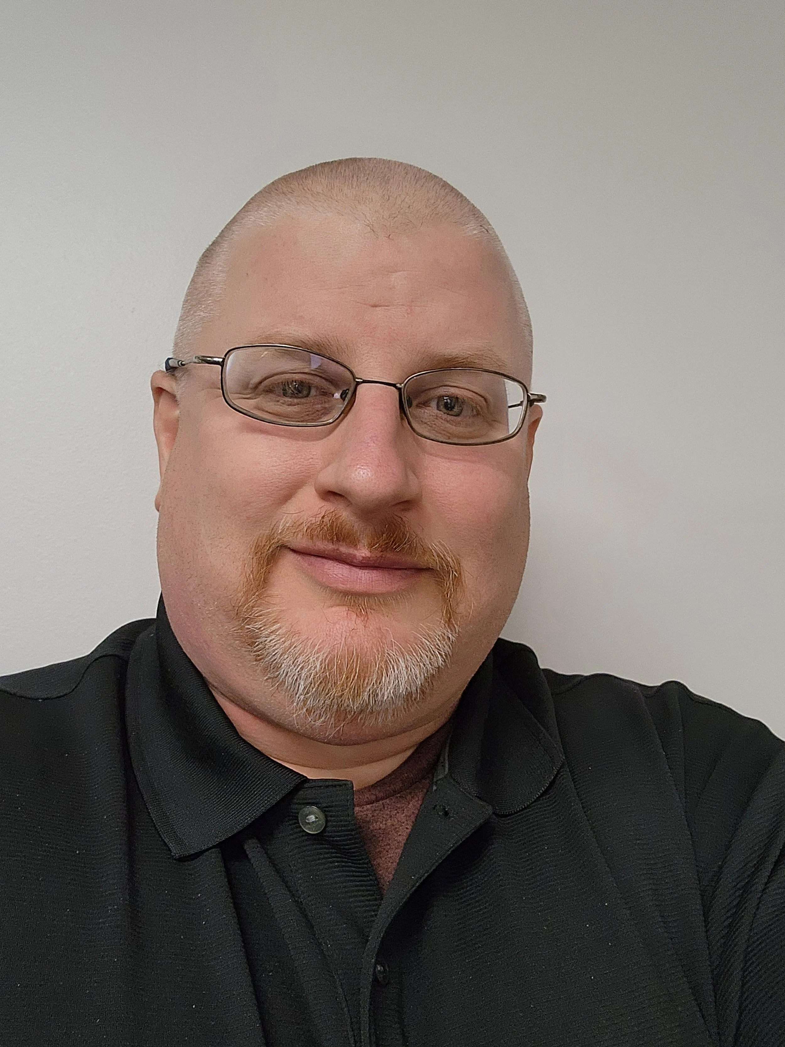 A man with glasses, a beard, and a shaved head, smiling, wearing a black collared shirt, taking a selfie against a plain white wall.