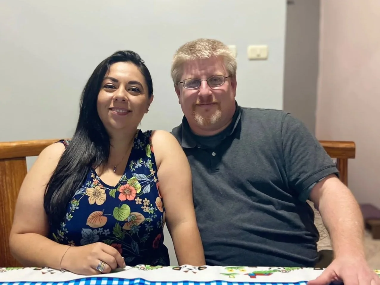 A woman with long dark hair and a man with glasses and blond hair sitting together at a table, both smiling at the camera.