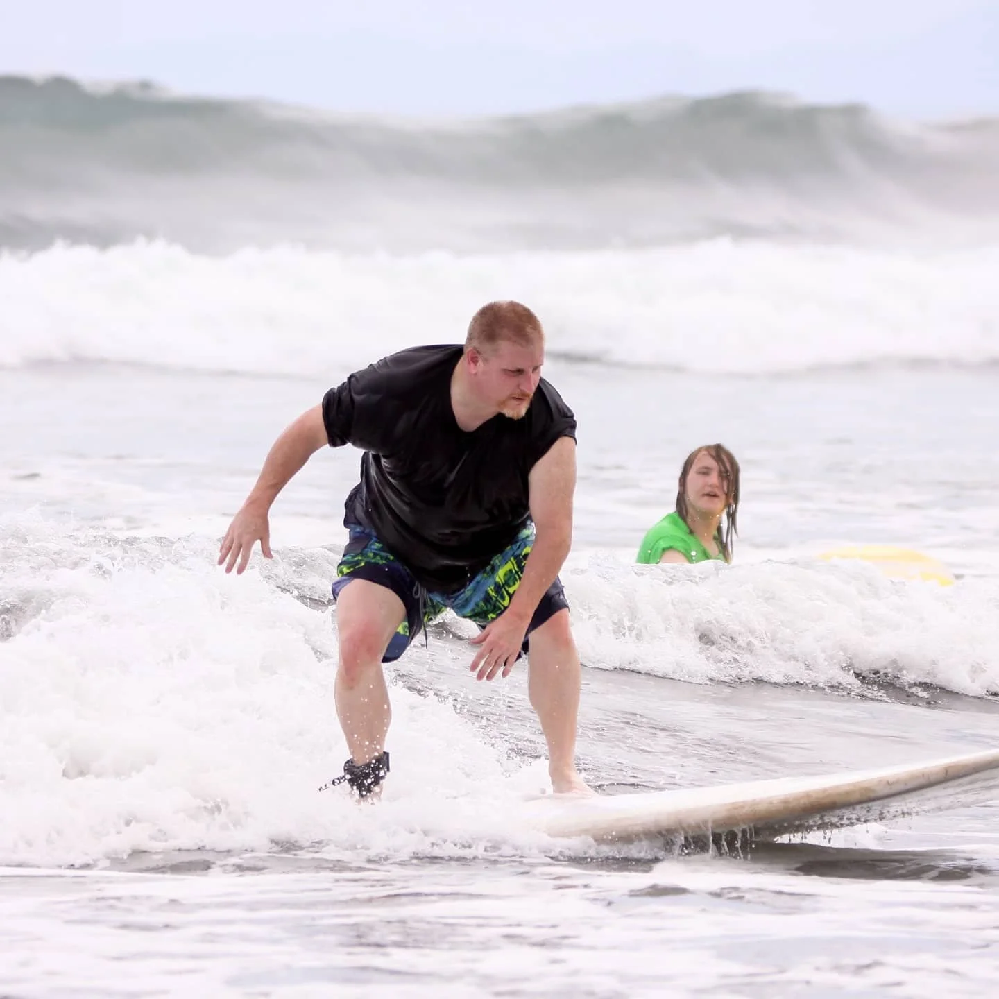 A man in a black T-shirt and colorful shorts attempting to surf on a small wave at the beach, with a boy in a green shirt in the background.