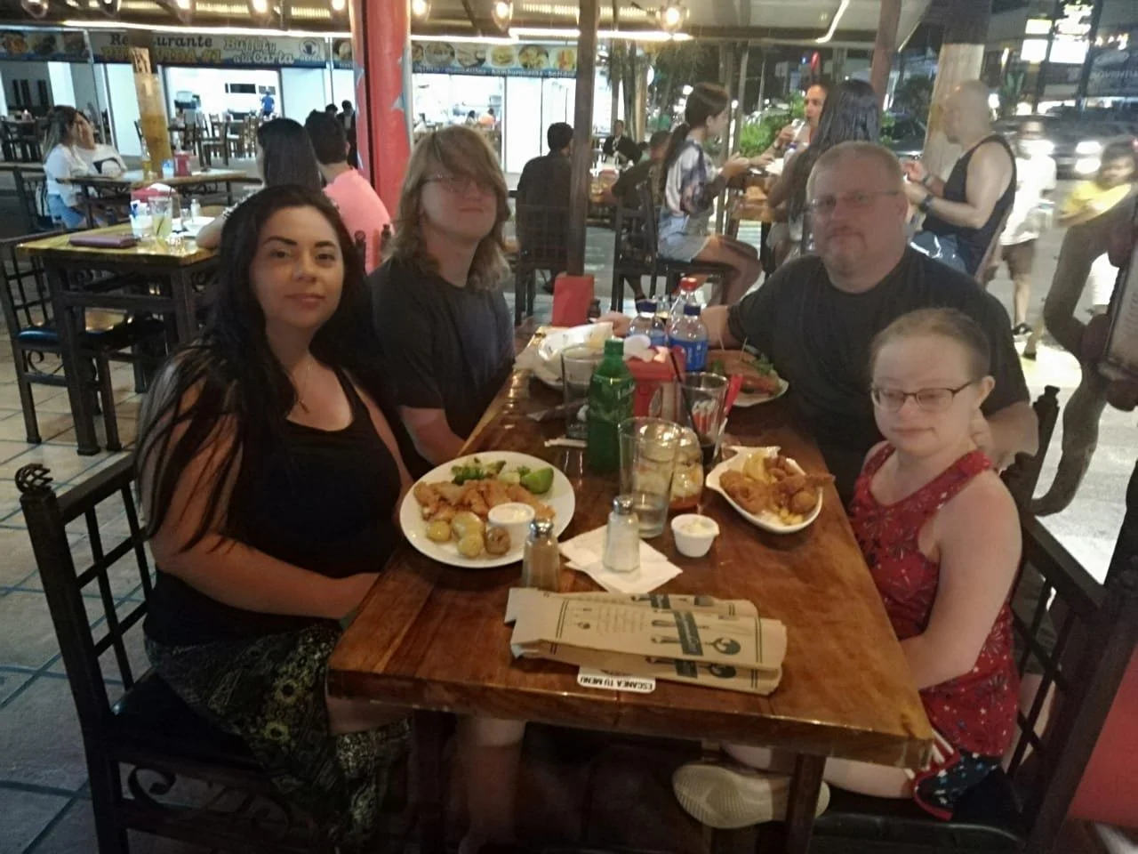 A family of four sitting at a restaurant table with food, indoors with other diners and some outside view visible through windows.