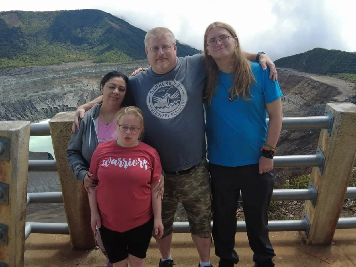 Group of five people standing in front of a volcanic crater with lush green mountains in the background. They are posing for a photo behind a railing, smiling and embracing each other.