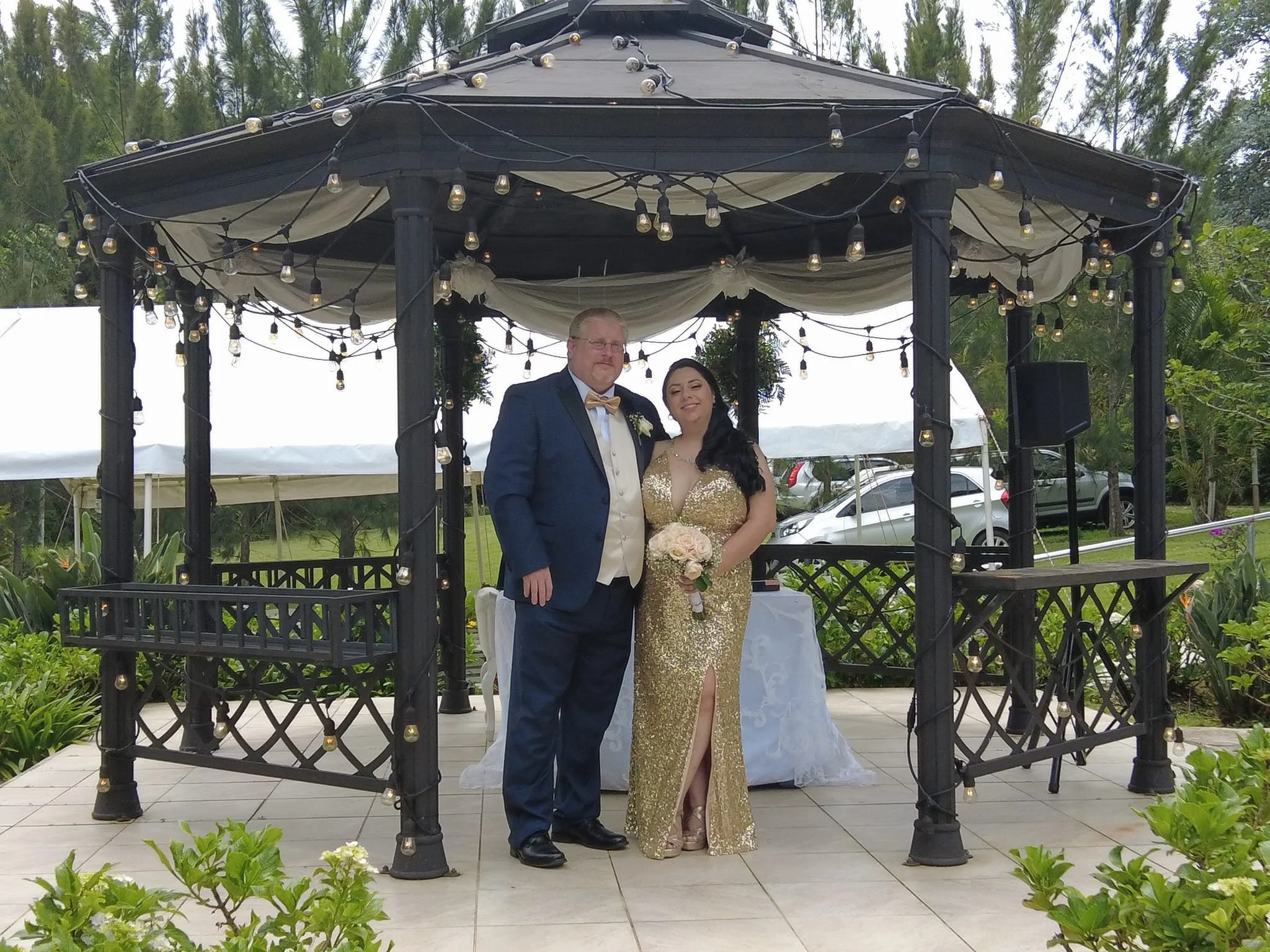 A newly married couple stands under a decorated gazebo outdoors. The groom wears a blue tuxedo with a cream vest and bow tie, while the bride wears a gold sequined gown with a slit, holding a bouquet of pastel roses.