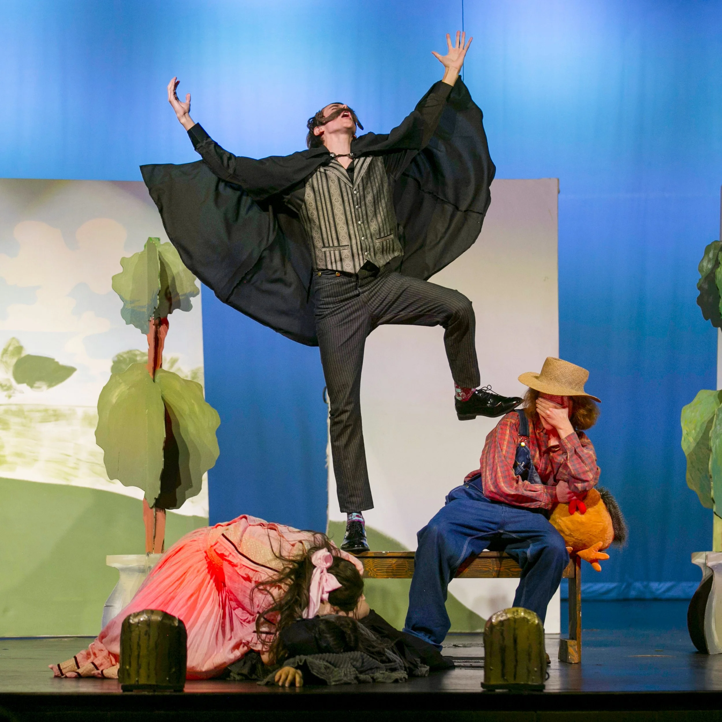 The scene is from a stage play with four actors. One person is standing on a bench, with one leg raised and arms outstretched, wearing a black cape and pinstripe pants. Another person is sitting on the bench in a plaid shirt and overalls, with a straw hat and a stuffed animal, covering their face. A third person is lying on the floor in a pink dress with a bow in her hair, covering her face with her hands. The background has painted scenery with trees and a blue sky.