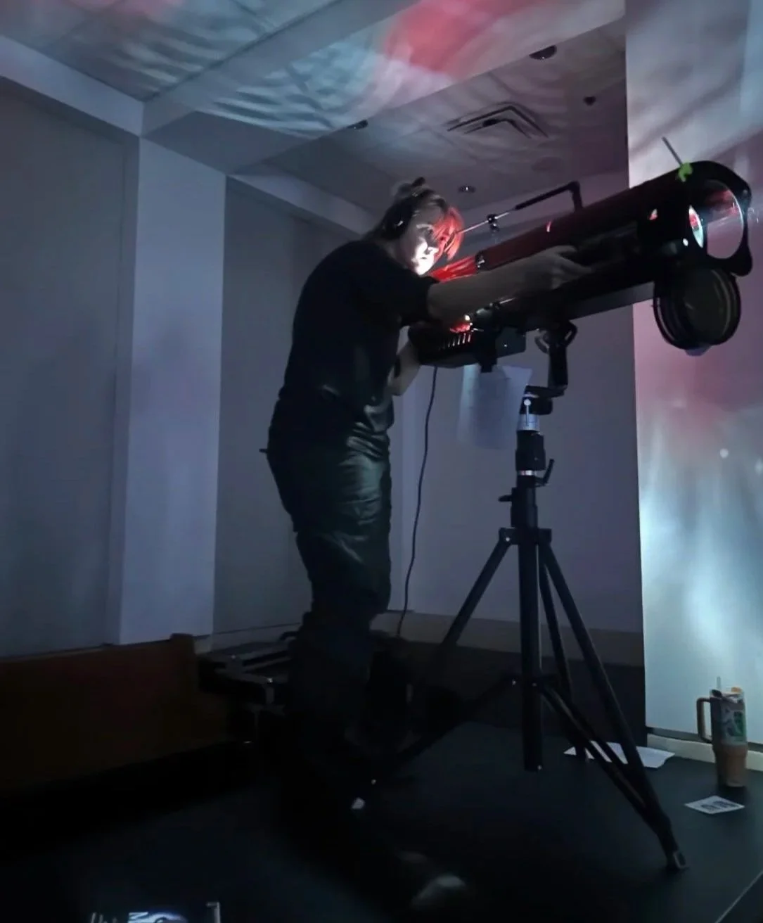 A person looking through a telescope mounted on a tripod in a dimly lit room.