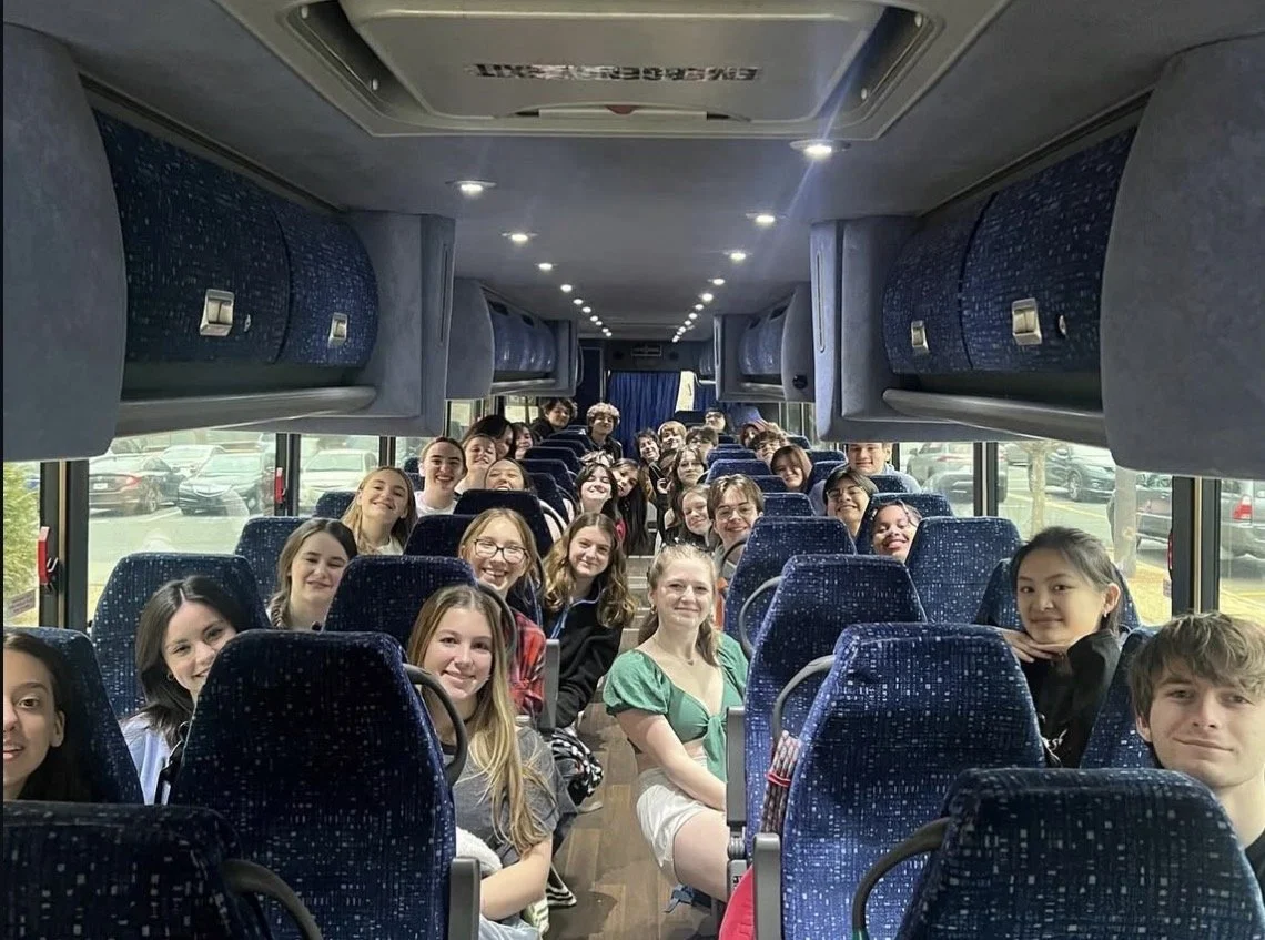 A group of people, mostly young women, sitting inside a bus with blue patterned seats, some facing forward and some turned to look at the camera, smiling and posing for a photo.