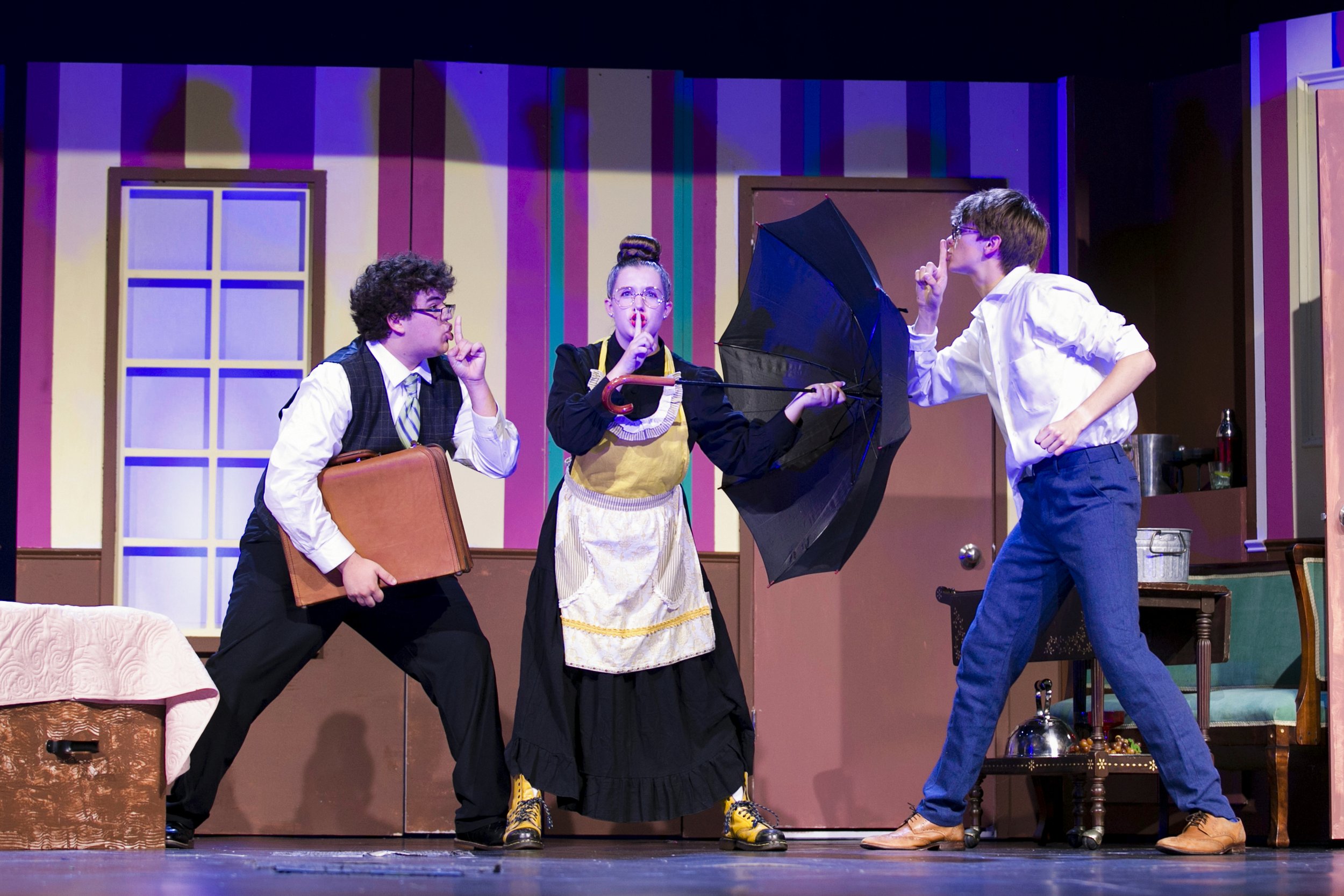 Three children performing a play on stage, with two boys and one girl, using props and costumes, in a colorful set with a couch and table.