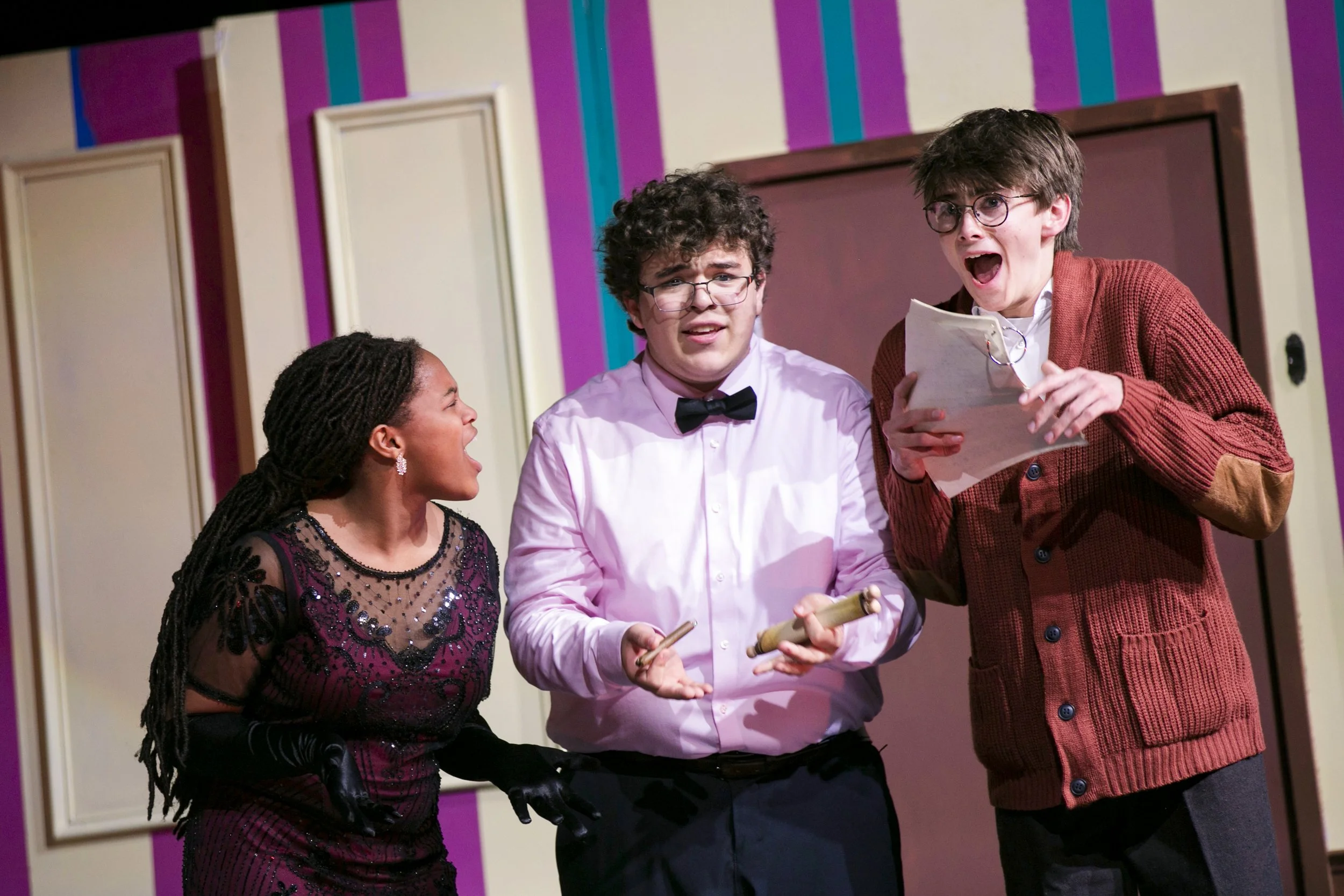 Three actors on stage, a woman on the left is talking passionately, a man in the middle with glasses and a bow tie looks confused, and another man on the right with glasses holds a script, appearing surprised or excited.