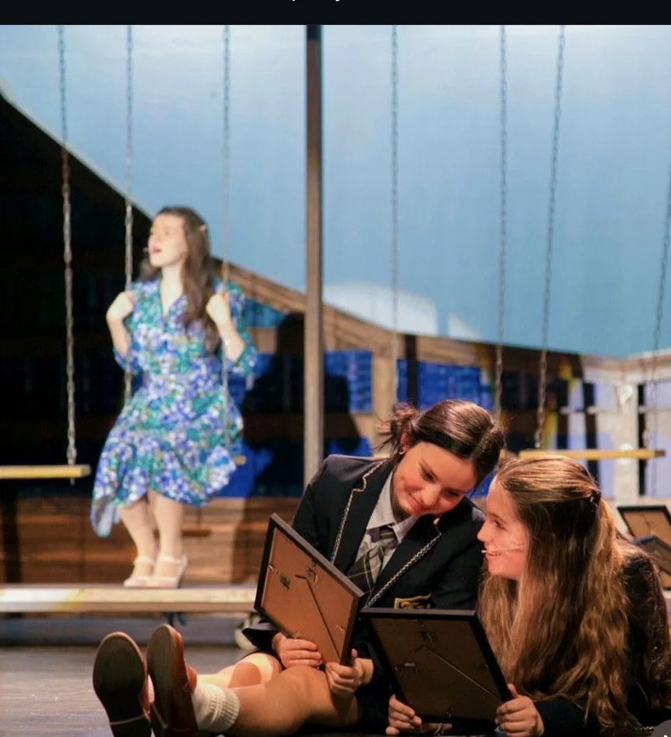 Two girls sitting on the floor reading framed pictures while a girl in a floral dress swings on a playground set in the background.