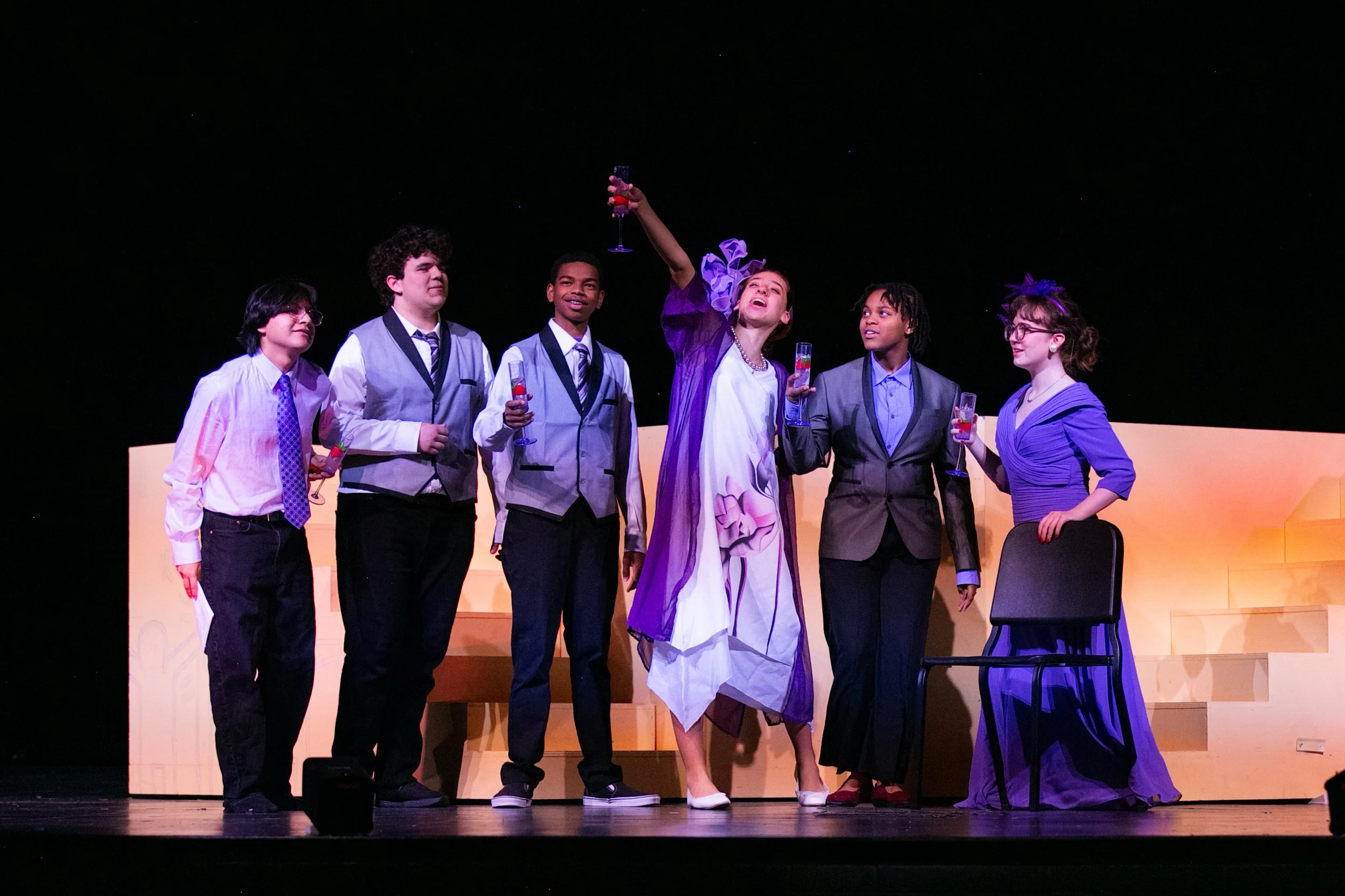 Six children in formal attire celebrating with drinks on stage during a theater performance.