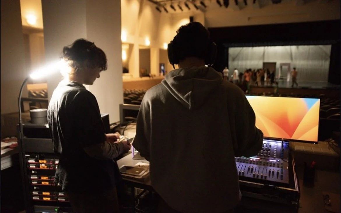 Two people wearing headphones working on audio equipment in a dimly lit theater or auditorium.