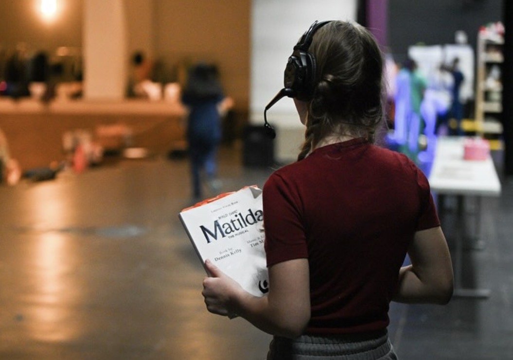 A girl wearing a maroon shirt and headphones, holding a script or newspaper titled 'Matilda,' standing in a theater or audition space.