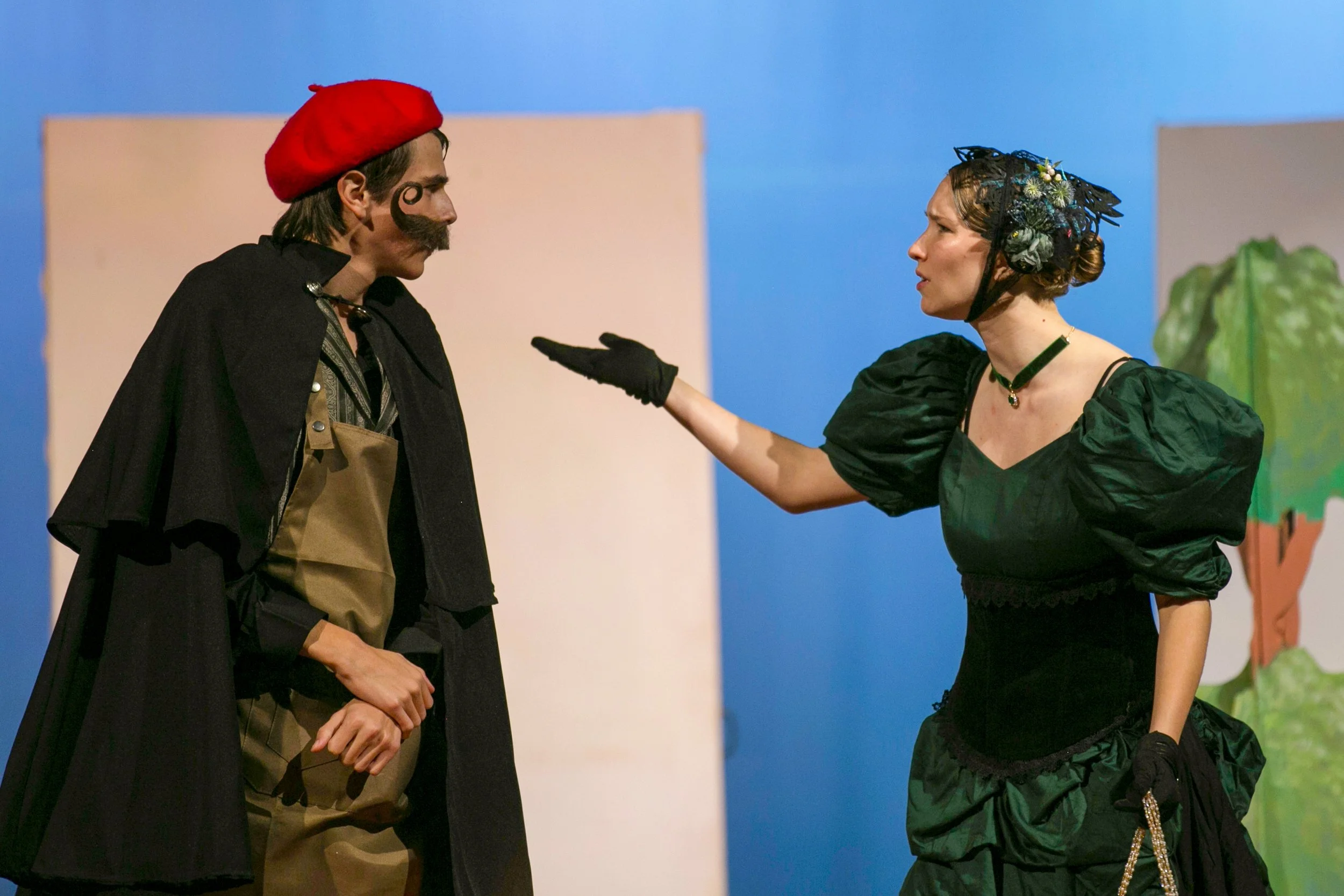 Two actors in costume performing on stage, with one woman pointing her finger at a man who appears to be listening intently.