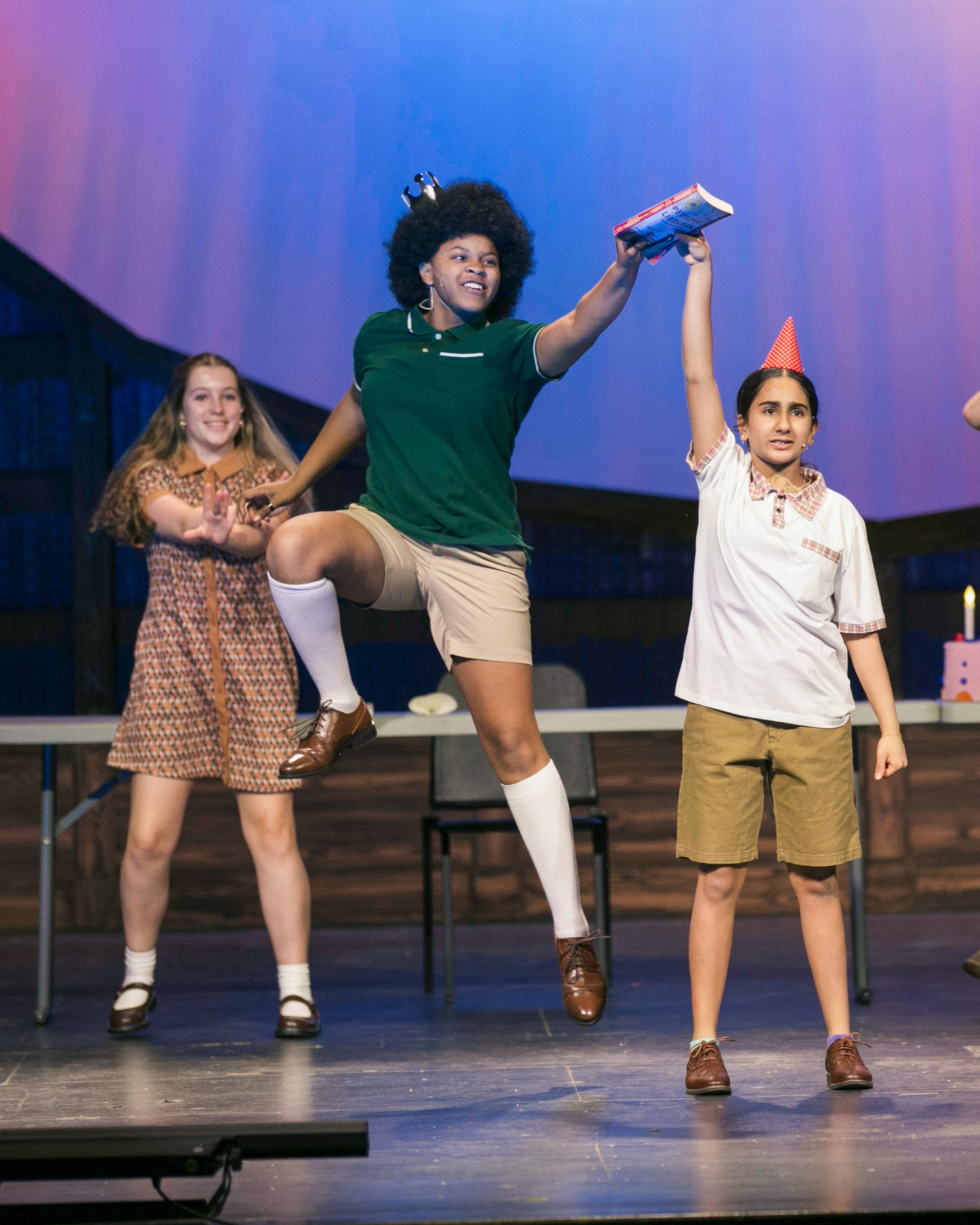 Three children performing on stage during a celebration, with one jumping with a book, another wearing a party hat, and a third clapping.