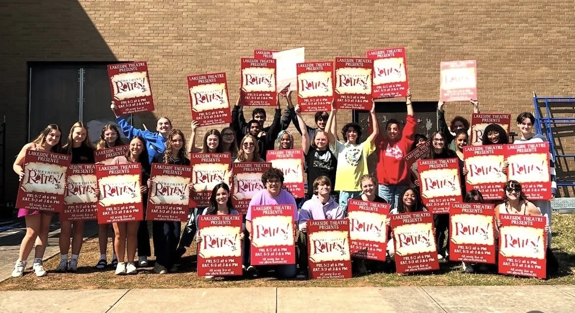 Group of young people standing outdoors in front of a brick building, holding signs with information about a theater production called "RENT!"