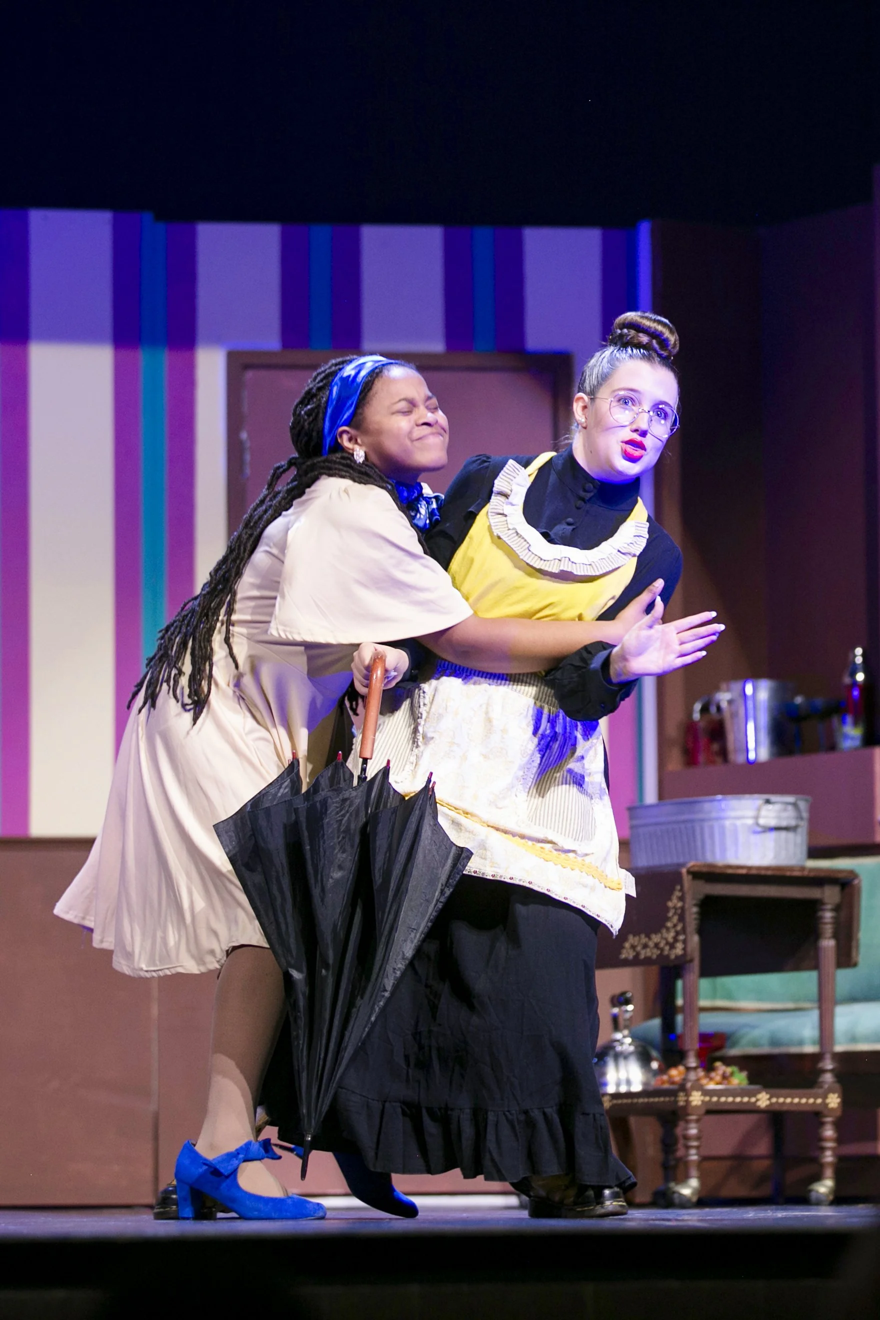 Two women perform on stage, one holding a black umbrella. One woman is dressed in a beige dress, blue shoes, and a blue headband, smiling with her eyes closed. The other woman is wearing glasses, a yellow apron, a black shirt, and a long black skirt, with her hair in a bun, and appears to be talking.