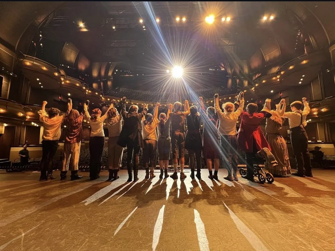 A group of people holding hands and raising them in the air on a stage in a theater, with stage lights shining down.