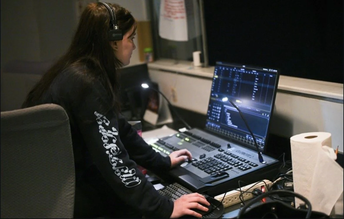 A woman working at a mixing console in a sound studio, wearing headphones, with a computer screen displaying audio editing software.