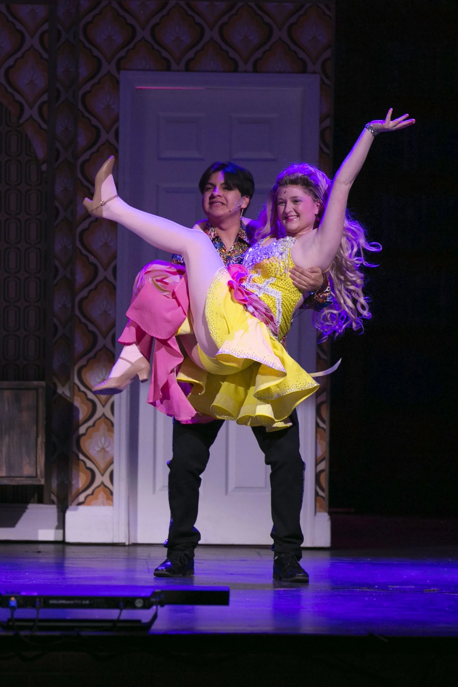 A man in black pants using his arms to support a woman in a bright yellow dress with curly blonde hair, who is lifting her leg high in a dance pose on stage with a vintage patterned backdrop.