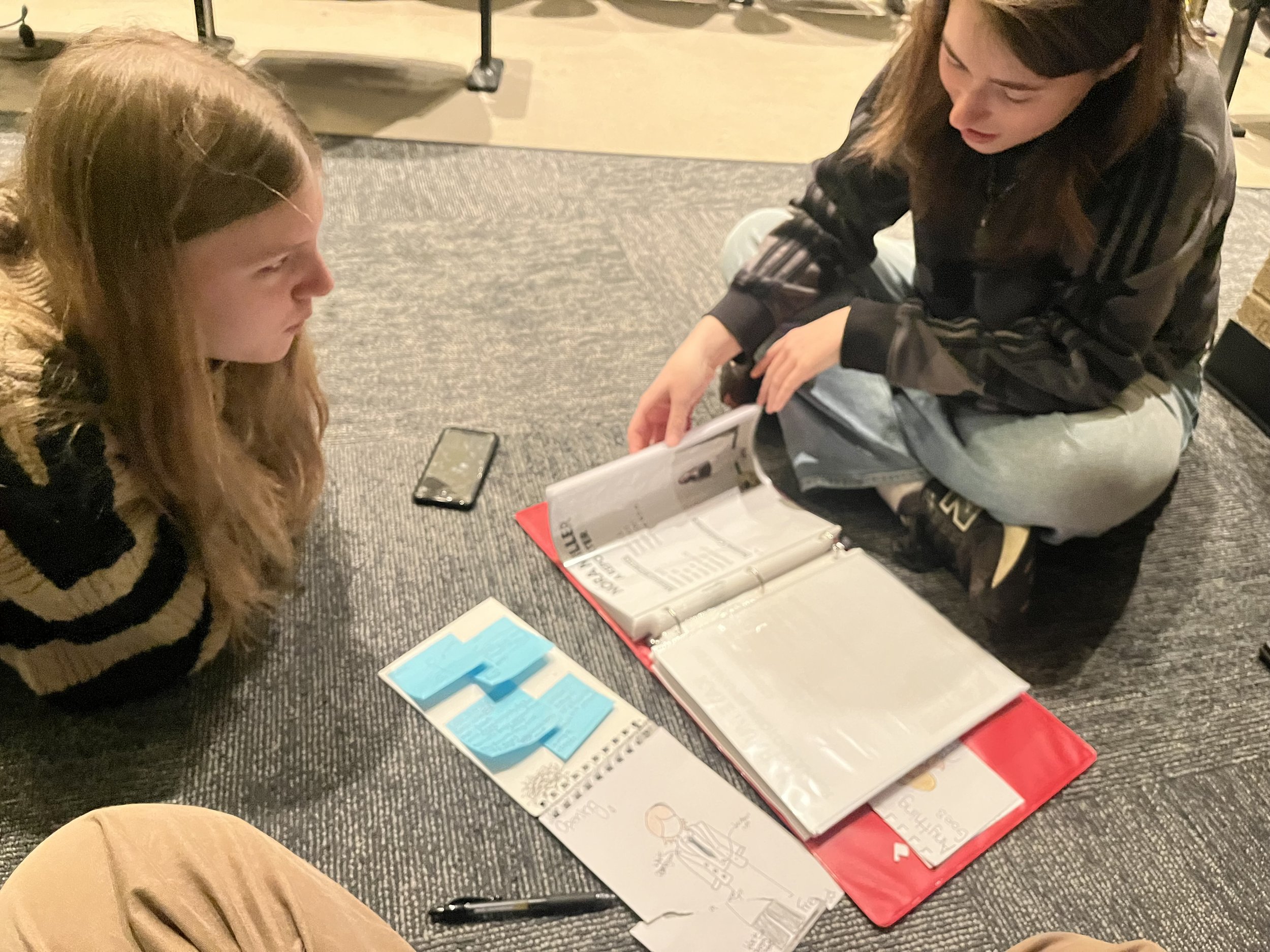 Two women sitting on a dark carpeted floor engaged in looking at a binder with papers, surrounded by sticky notes, a notebook, and a pen. One woman has long red hair and is wearing a striped sweater, while the other has brown hair and is wearing a black and gray jacket.