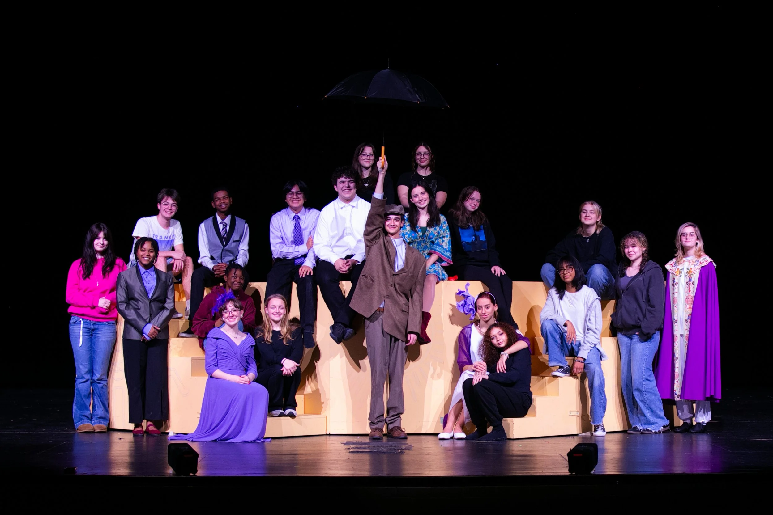 A group of young people on a stage, some sitting on a multi-level beige platform, with one person standing in front holding an umbrella. The stage background is black, and the group is dressed in casual and semi-formal clothing, smiling at the camera