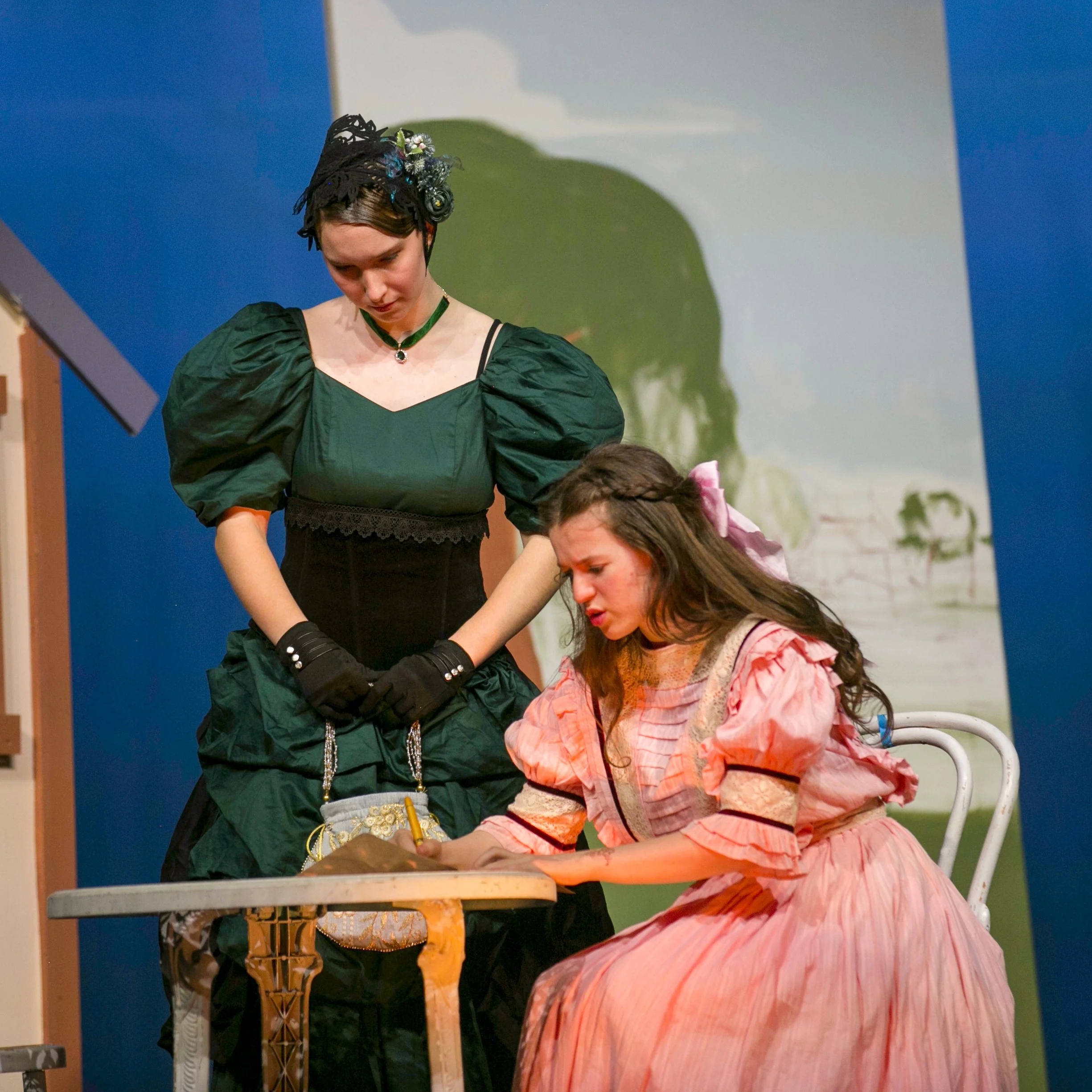 Two women in period costumes on stage, one sitting at a table and writing, the other standing beside her, overseing the writing, with a landscape painting in the background.