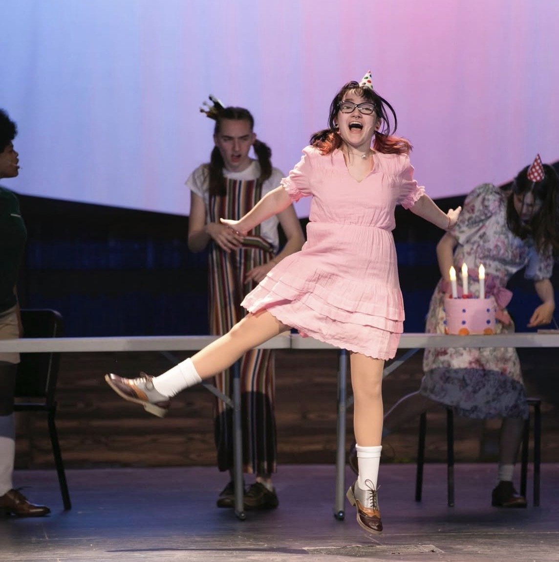 A girl in a pink dress is jumping and smiling during a birthday celebration with a cake and candles in the background.