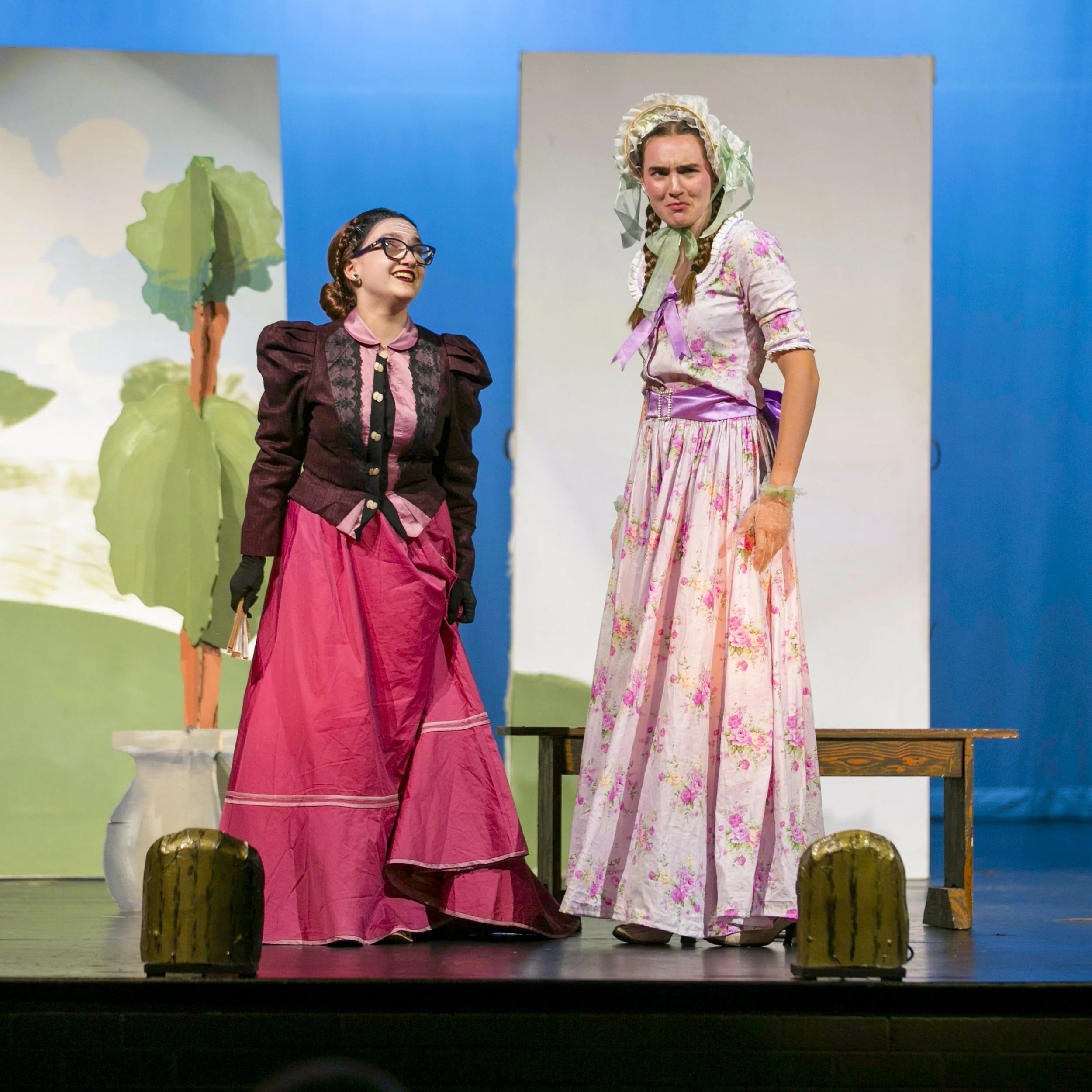 Two women on stage in period costume, one smiling and one looking skeptical, with a painted backdrop of trees behind them.