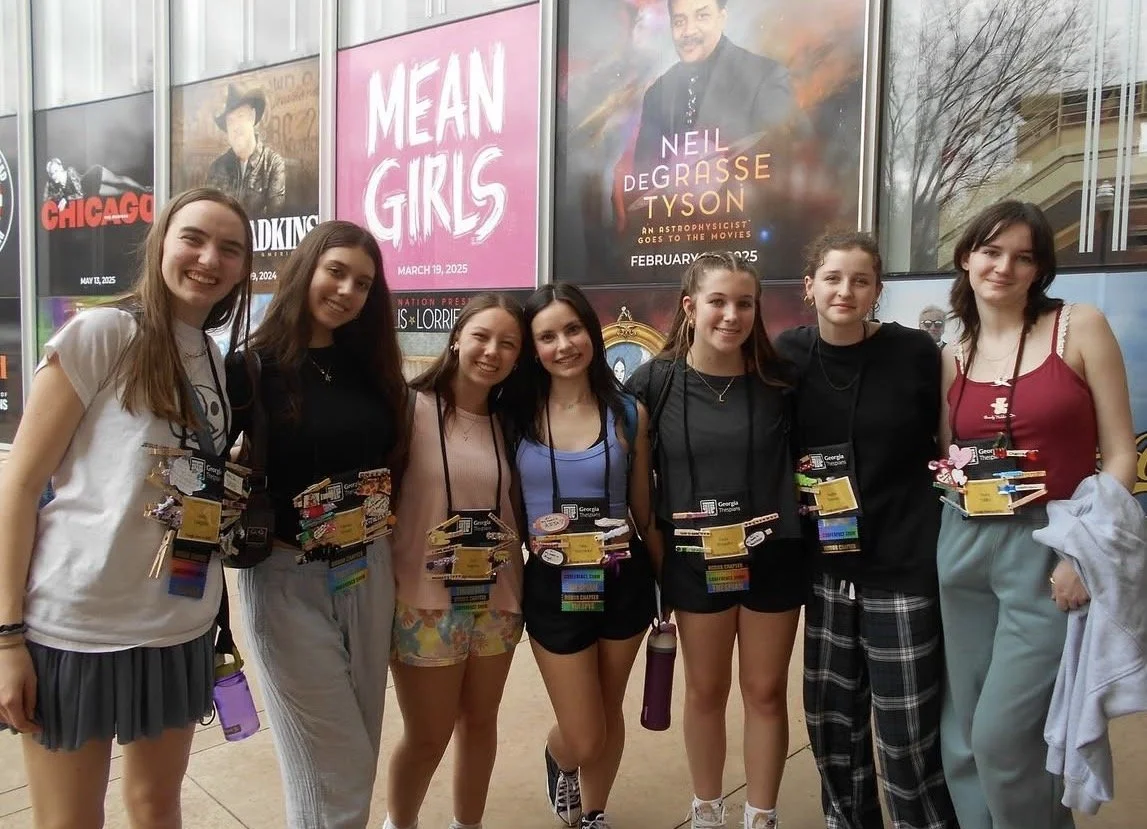 A group of seven young girls standing together inside a building, smiling at the camera. They are wearing casual clothes and badge necklaces, and there are posters for Broadway shows behind them, including 'Mean Girls' and others.