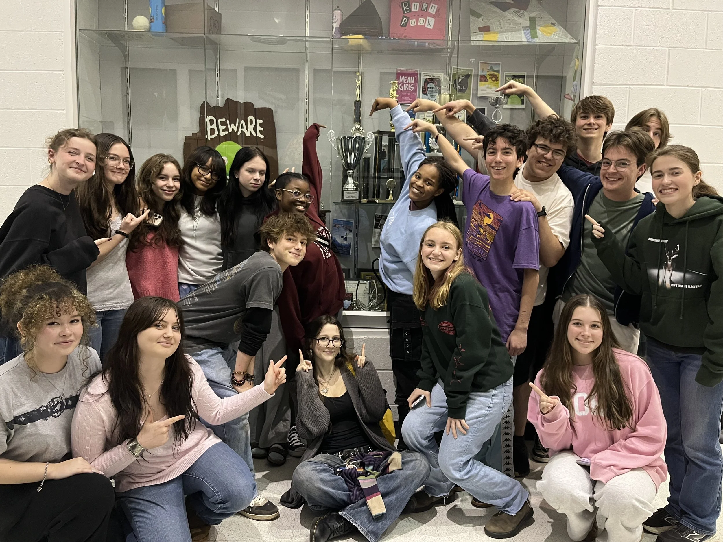 A group of teenagers and a woman posing in front of a display case with trophies and a 'Beware' sign, some pointing and smiling at the camera.