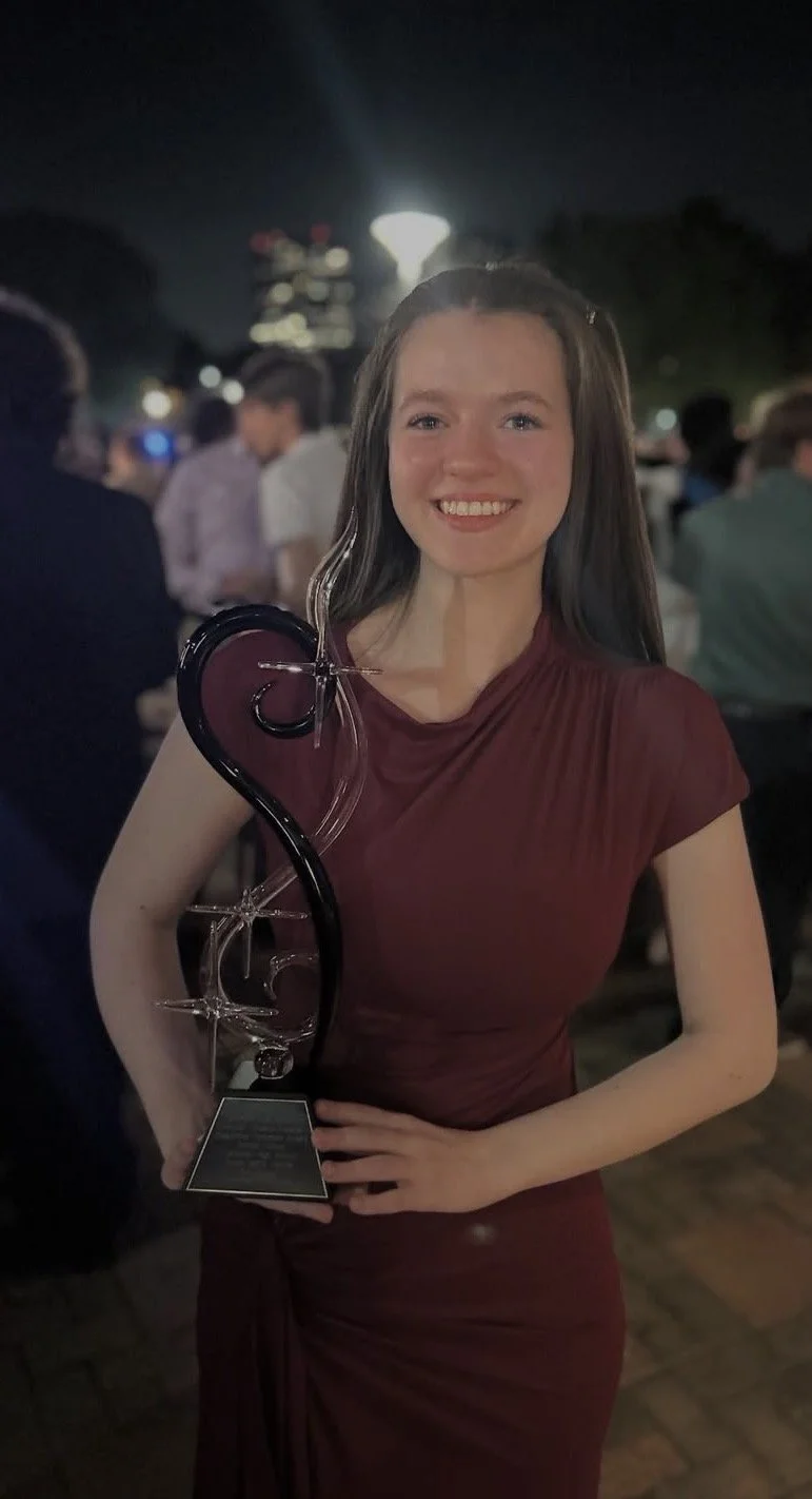 A young woman wearing a dark red dress smiling at night while holding a glass award trophy.