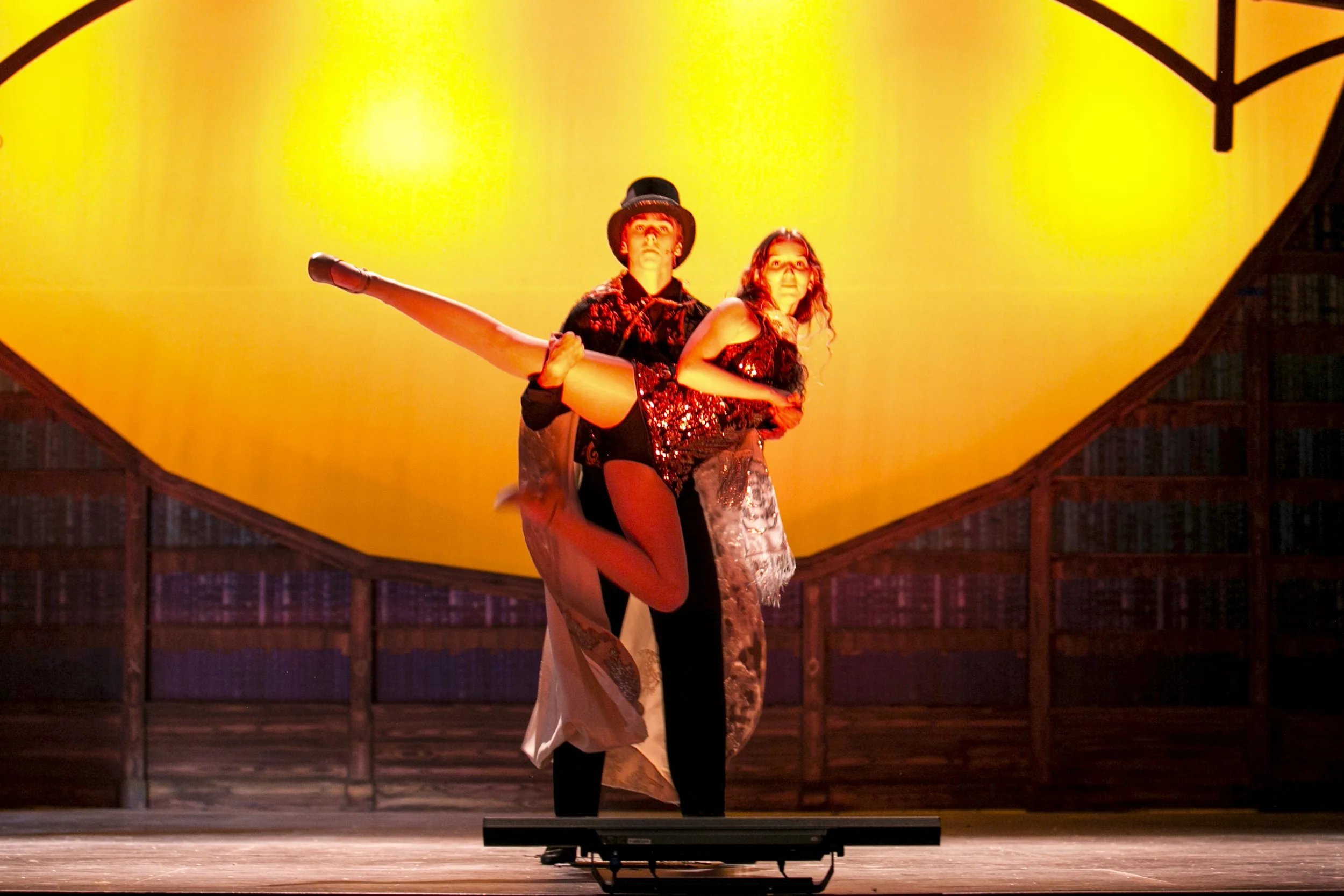 A man and woman performing a dance on stage. The man is holding the woman in a pose, with one leg lifted and extended. They are dressed in theatrical costumes, with a bright yellow backdrop.