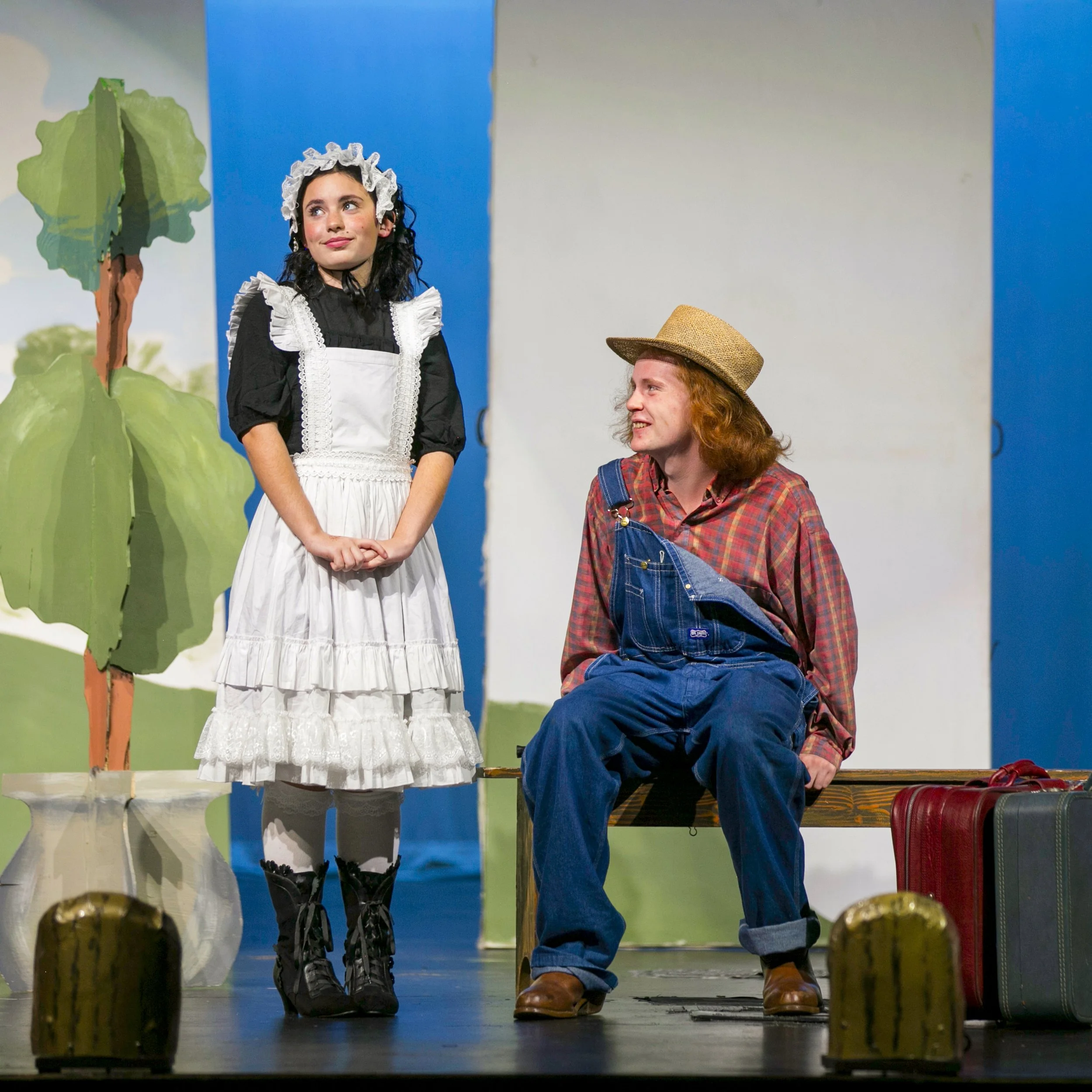 Two actors on a stage, one dressed as a young girl in a black dress, white apron, and bonnet, and the other as a man in overalls, plaid shirt, and straw hat, with a painted backdrop of trees and sky.