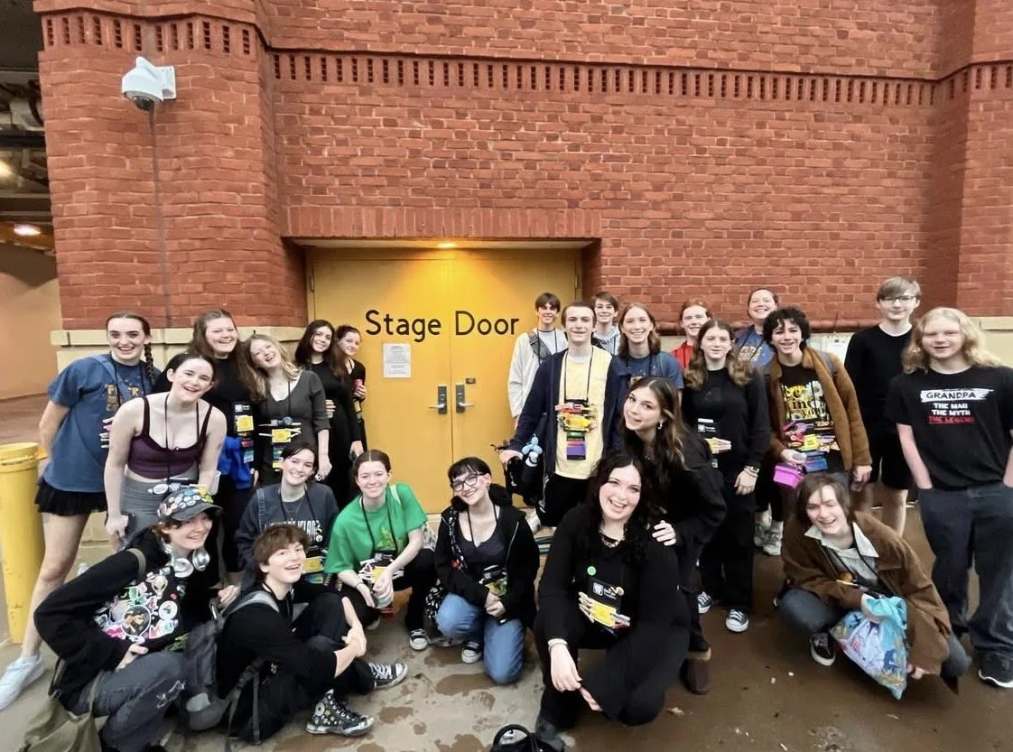 Group of young adolescents, mostly girls, posing in front of a yellow door labeled 'Stage Door' under a brick building. They are smiling, wearing casual clothes, and some have backpacks or headphones.