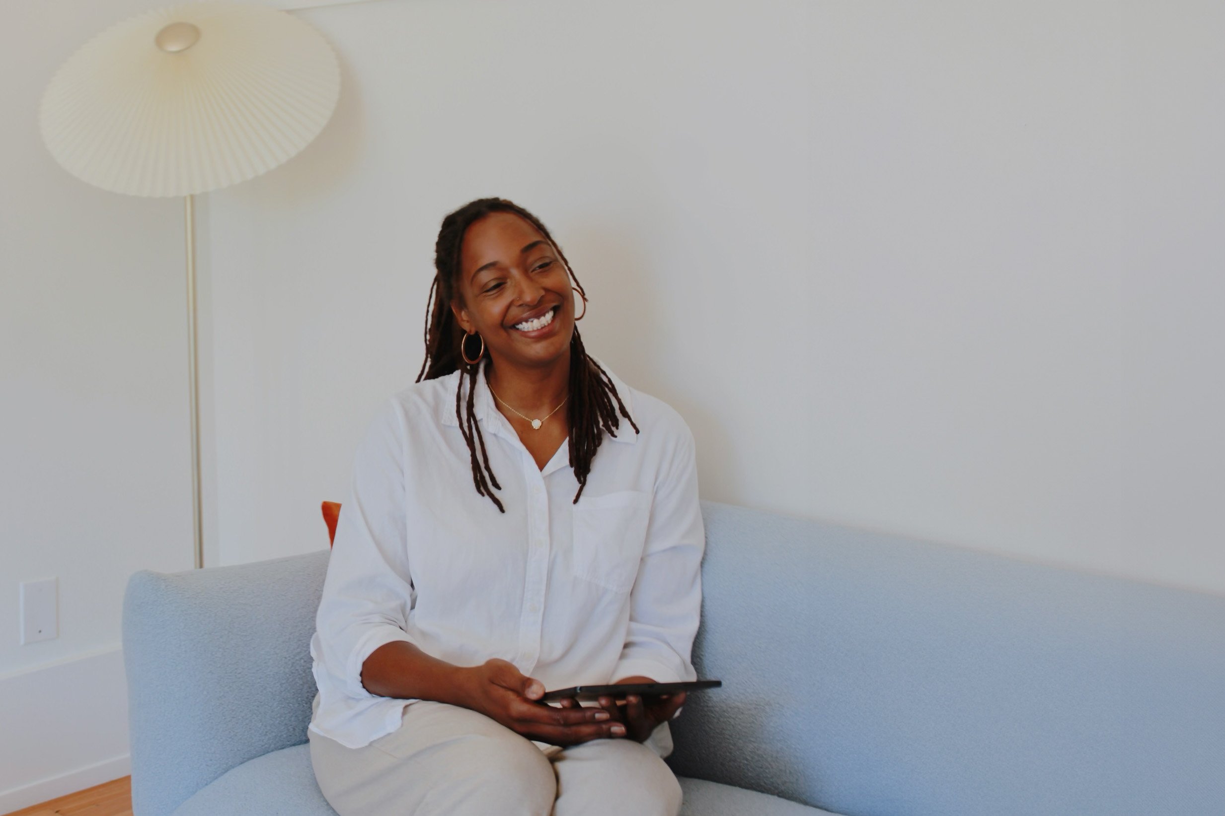 A woman with dreadlocks and a white shirt sitting on a light blue sofa, holding a tablet, smiling, with a white wall and a floor lamp behind her.