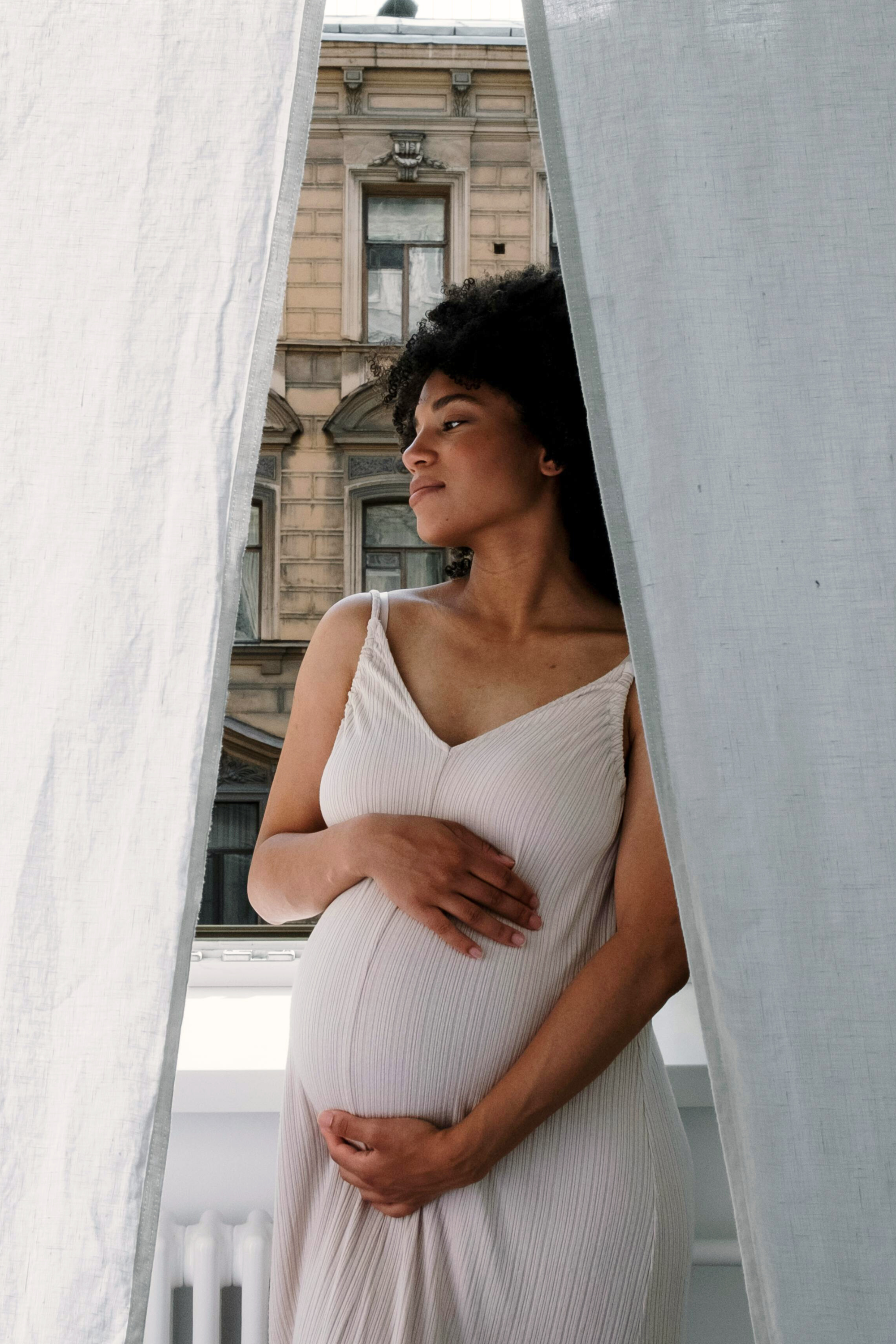 A pregnant woman in a beige, ribbed dress stands between white curtains, looking thoughtfully out a window at a building with ornate architecture.
